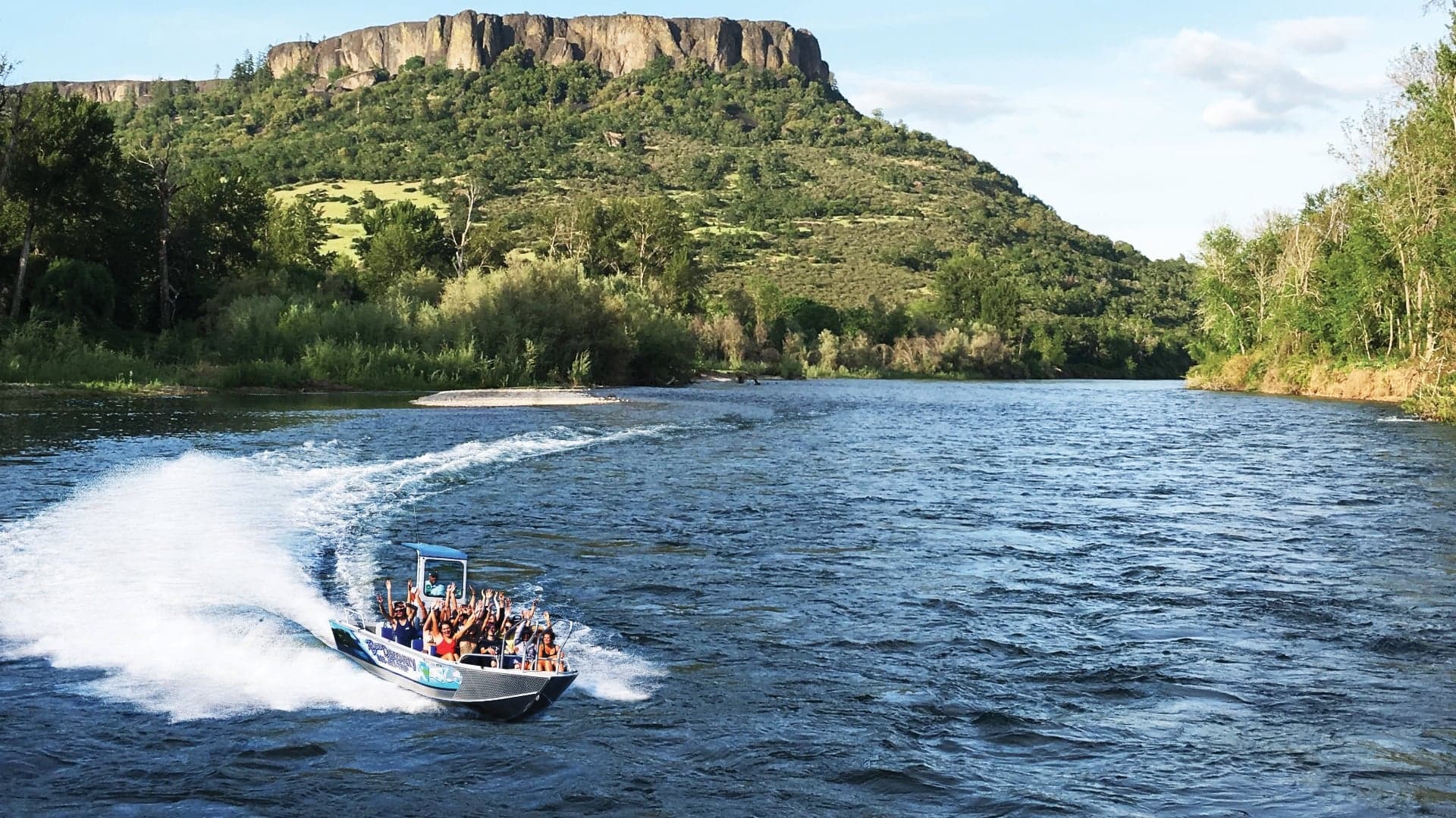 A speedboat with many passengers speeds across a river, surrounded by lush greenery and a rocky hill in the background.