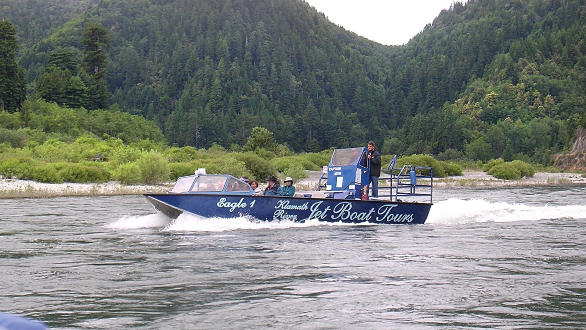 A blue jet boat named "Eagle 1" navigates the Klamath River, surrounded by lush green mountains.