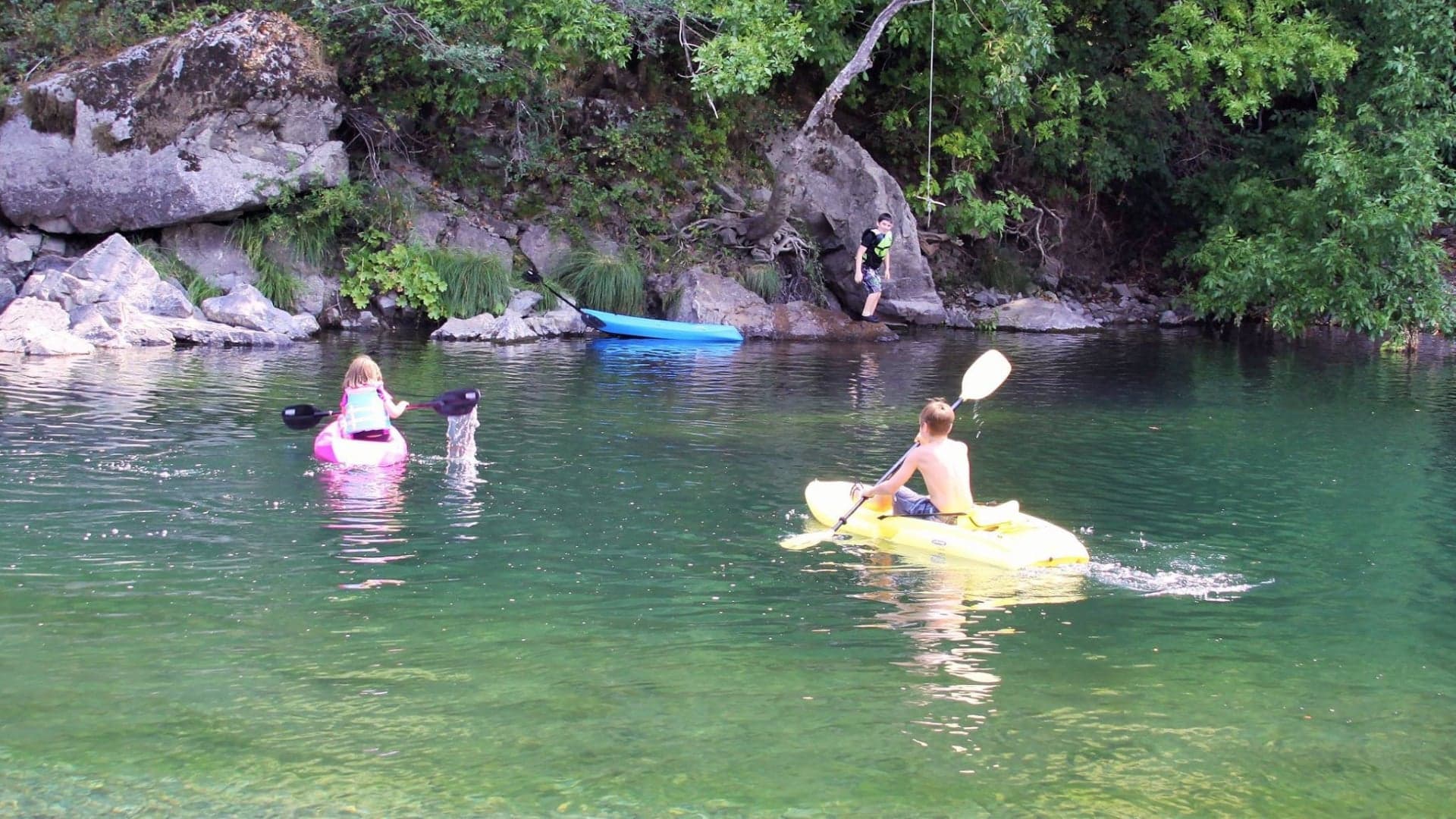 Two children kayak on a river surrounded by greenery, with rocks and a blue kayak visible nearby.