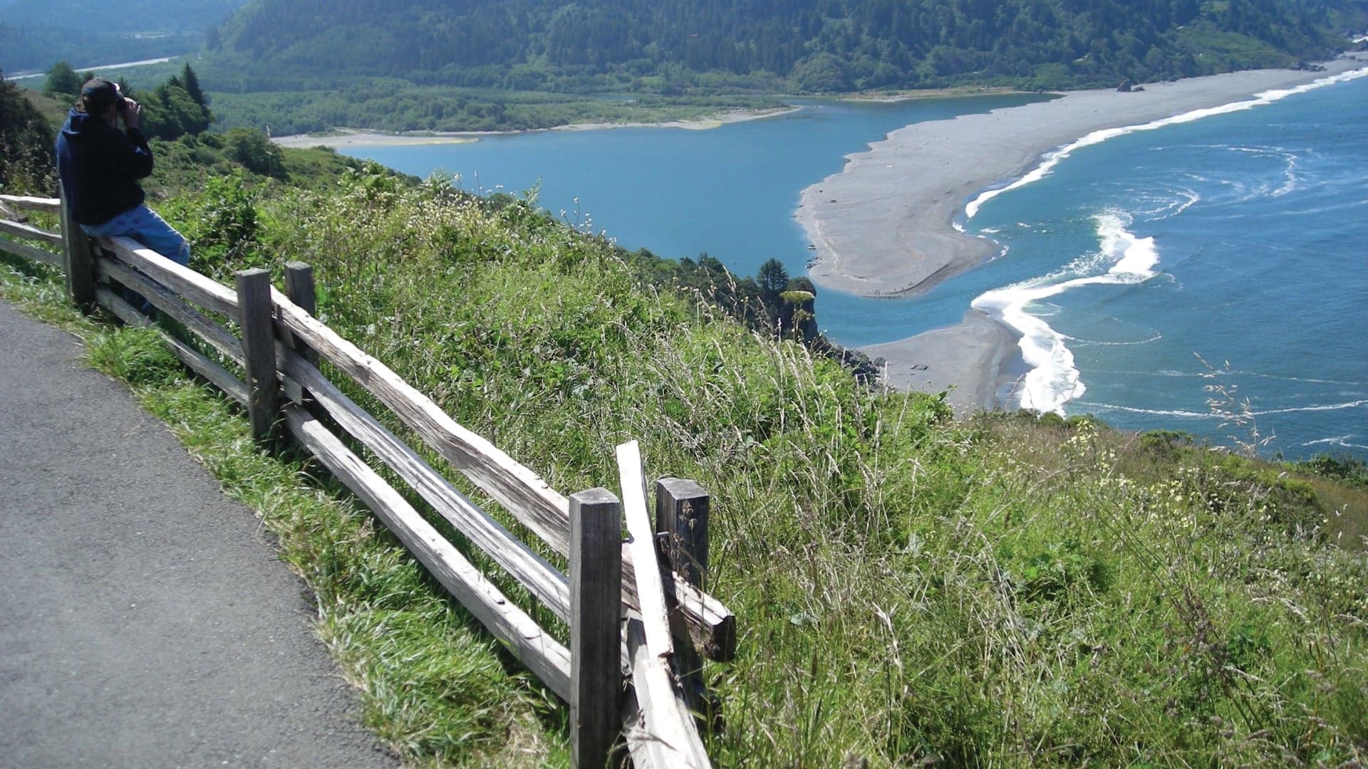 A person sits on a wooden fence, overlooking a river and beach surrounded by green hills under a clear sky.
