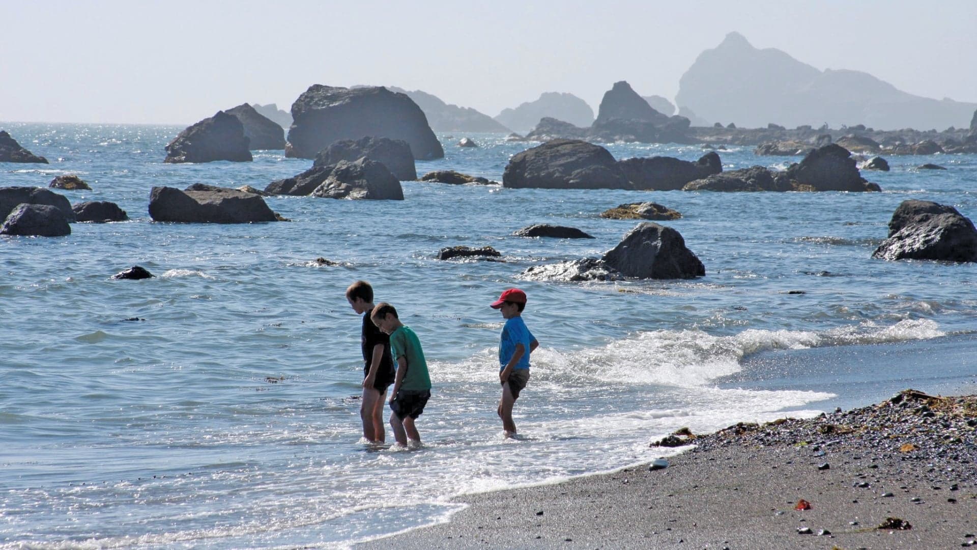 Three children wade in the shallow water at a beach, surrounded by rocks and gentle waves.