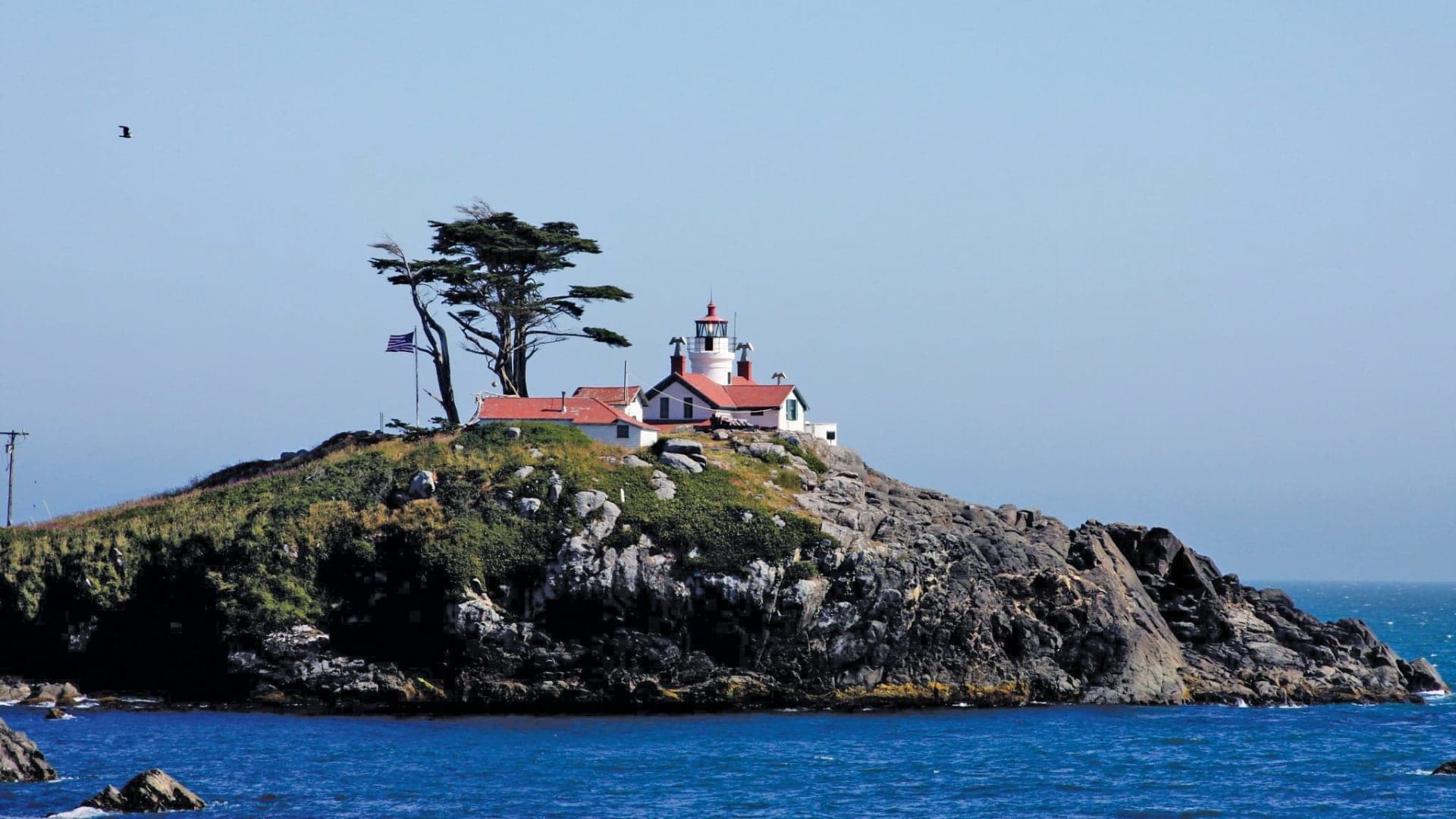A lighthouse on a rocky island, surrounded by blue ocean and a clear sky, with a flag and trees nearby.