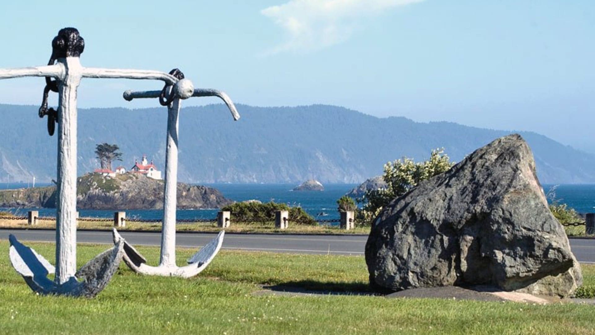 Two large anchors in a grassy area, with a coastal view of an island and distant mountains under a clear sky.