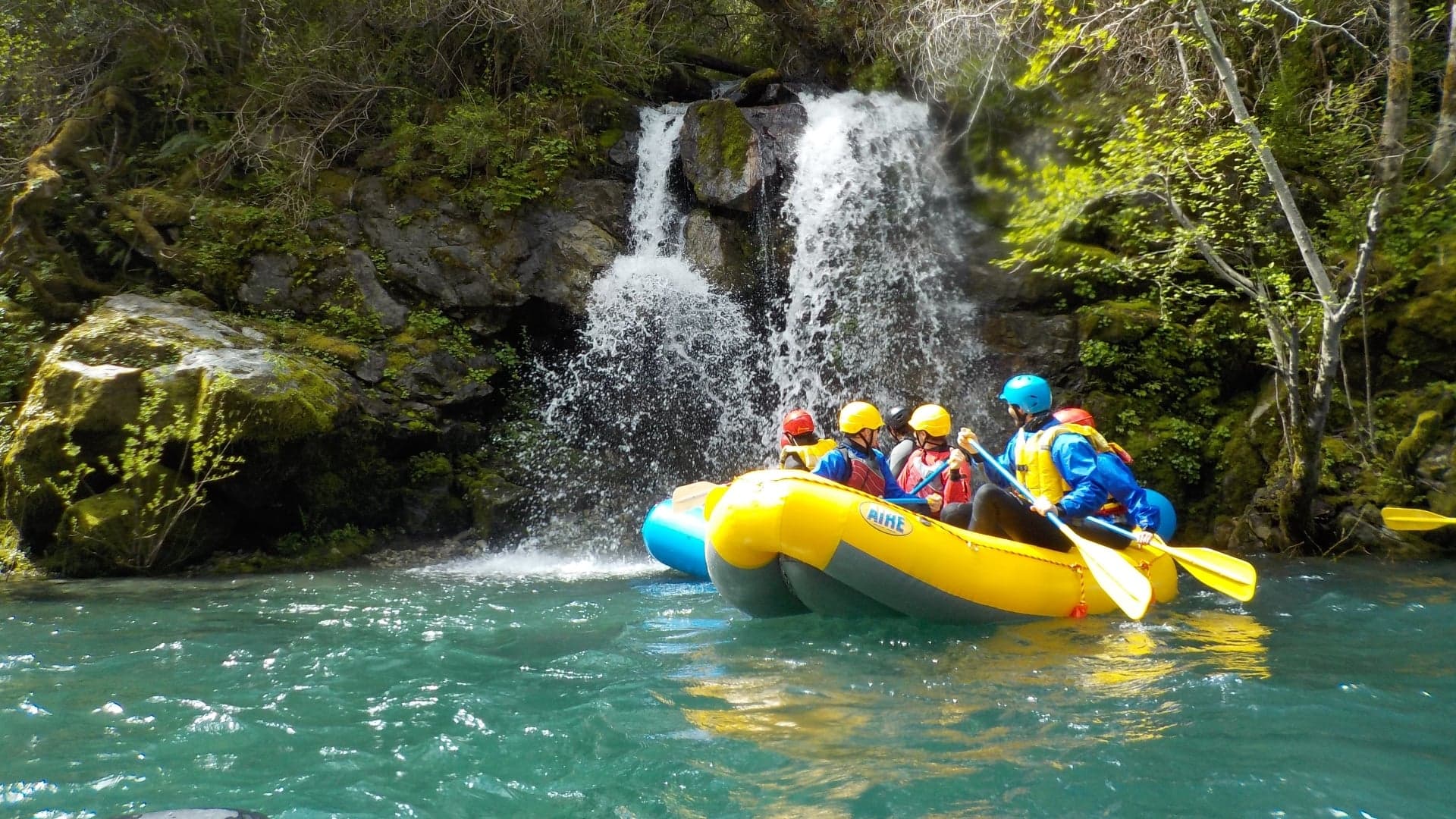 A yellow raft with four paddlers approaches a small waterfall surrounded by lush greenery.