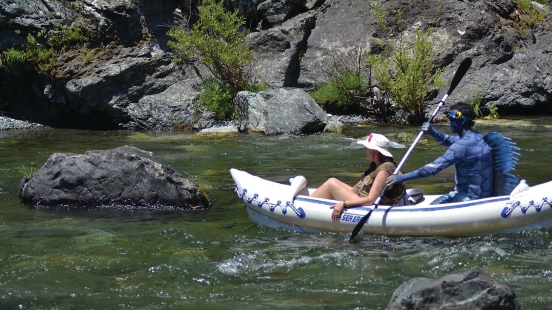 A woman in a kayak with a blue, humanoid creature (costume) paddling along a rocky, shallow river.