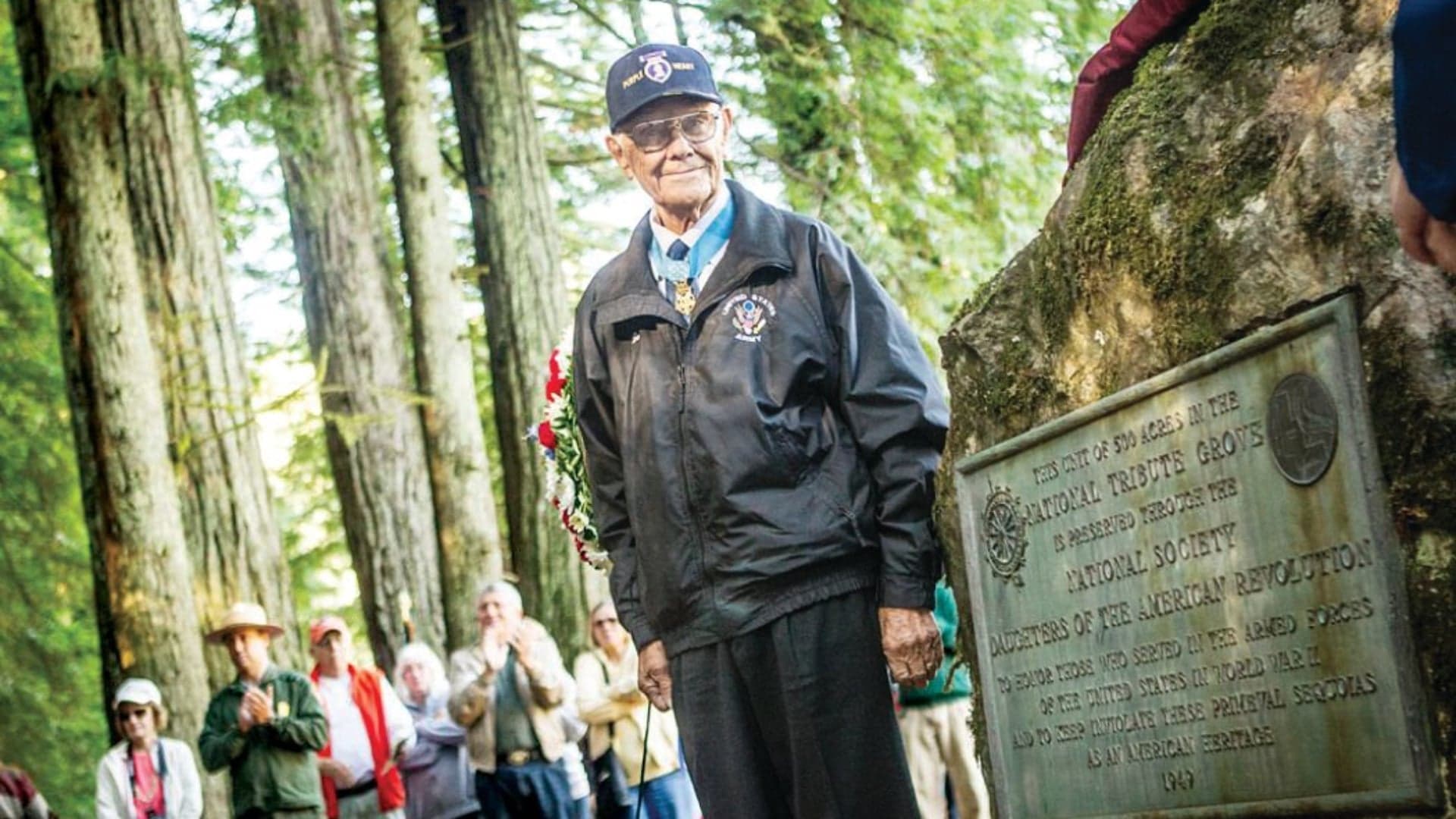 An elderly man in a jacket and cap stands near a memorial plaque, surrounded by an audience in a forest.