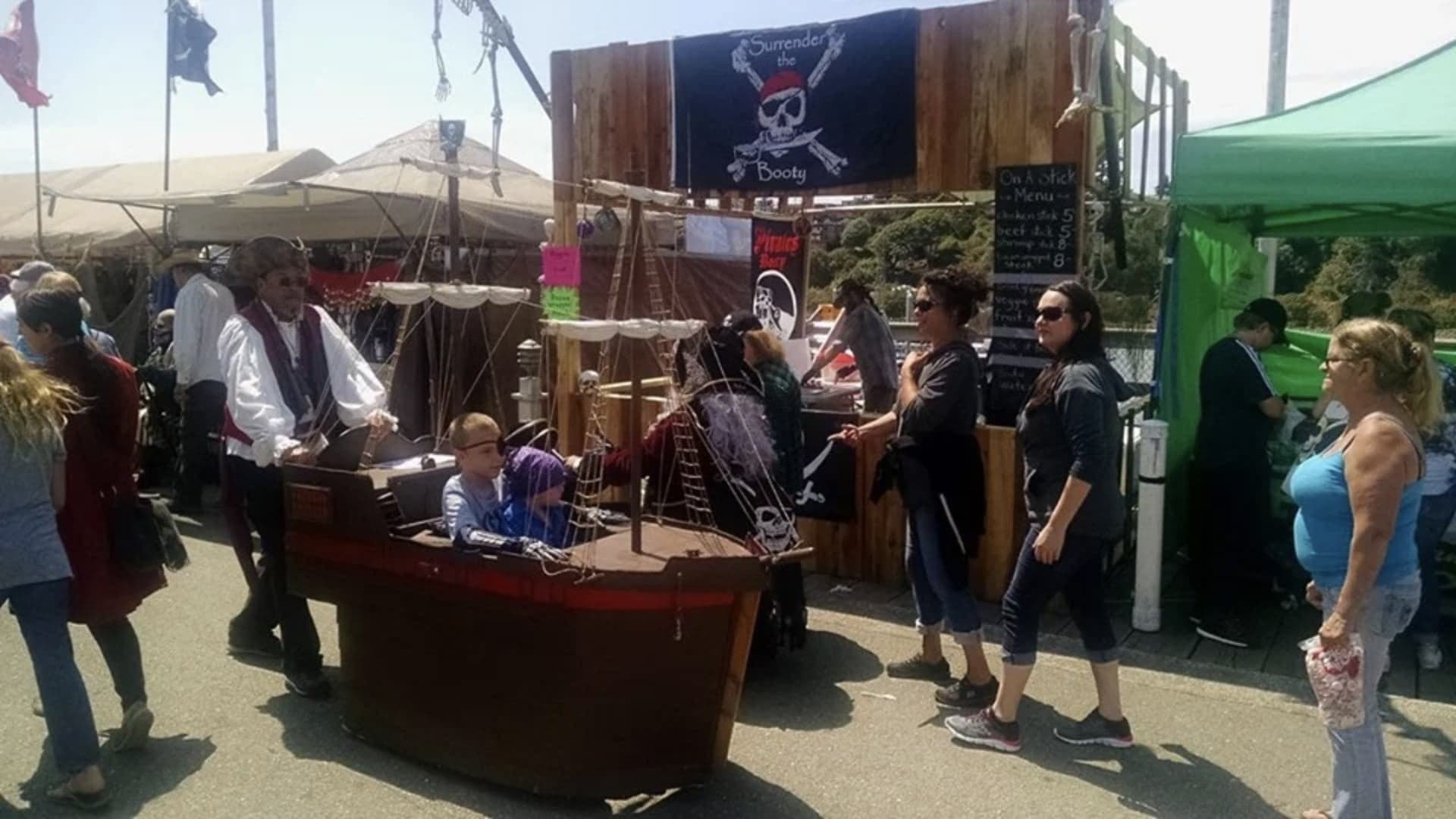 A pirate-themed booth at an outdoor event with people interacting, and a child sitting in a model pirate ship.