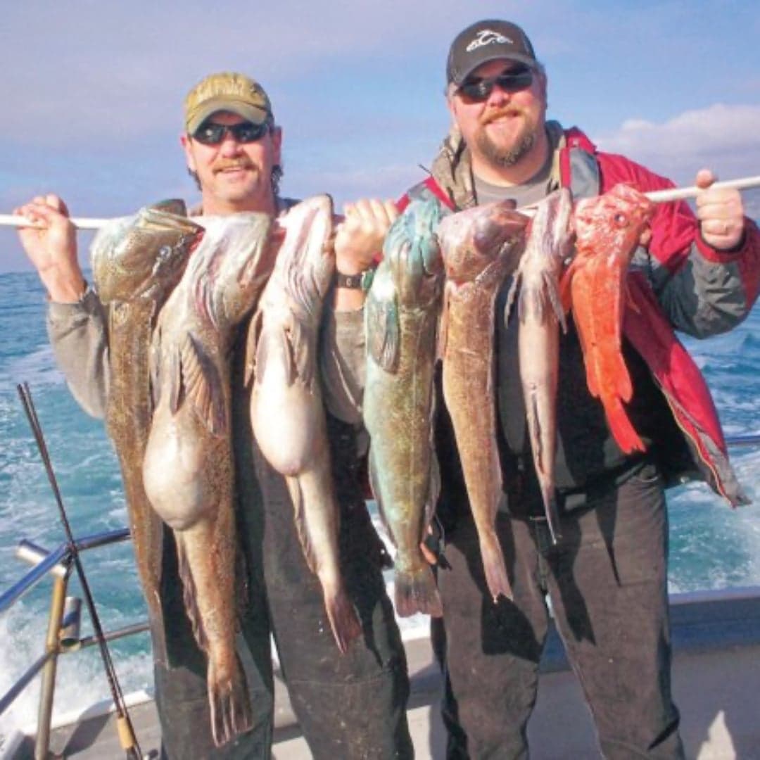 Two men hold a large catch of fish on a fishing boat, smiling at the camera.