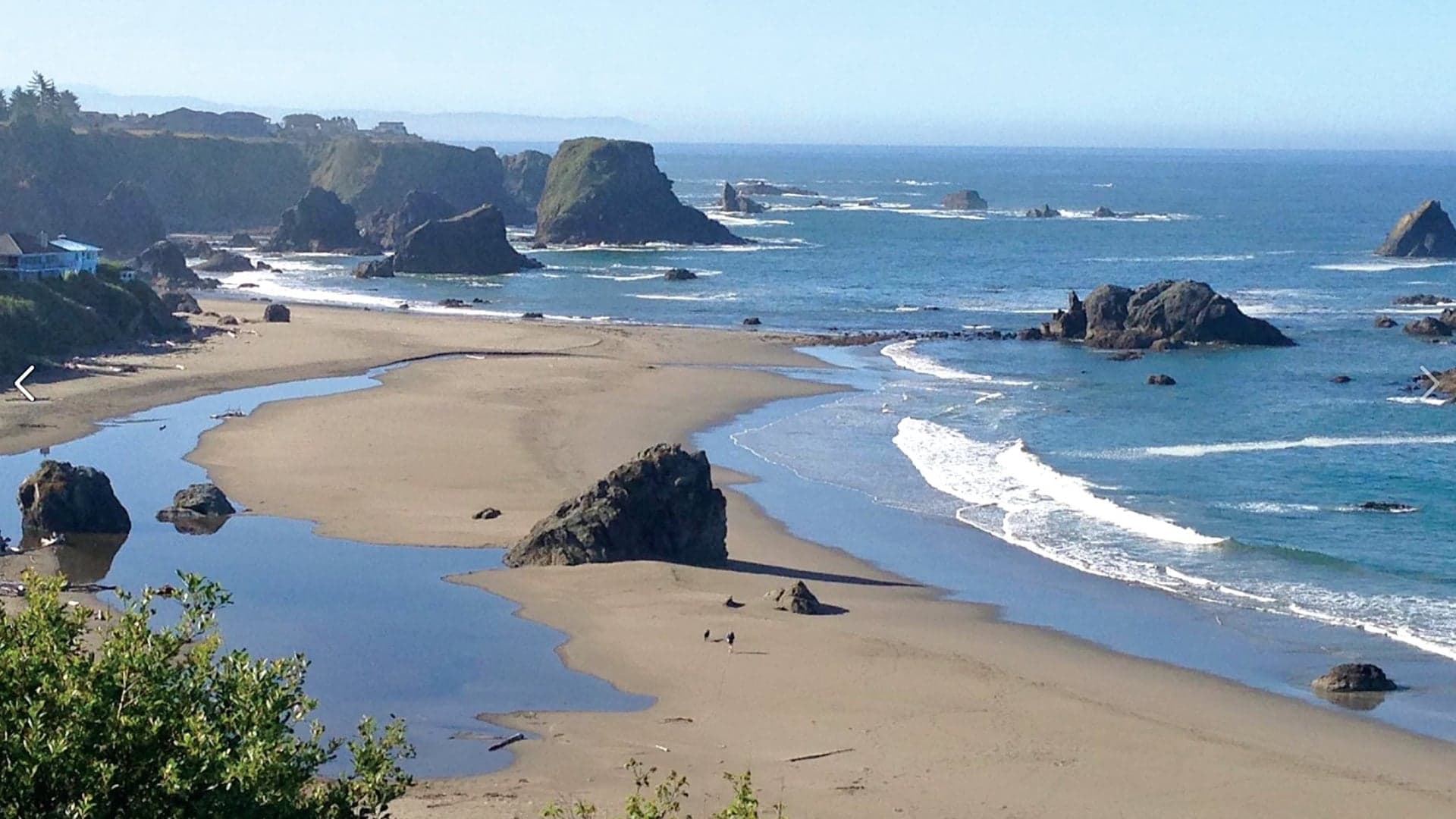 Rocky coastline with sandy beach, tidal pools, and gentle waves under a clear blue sky.