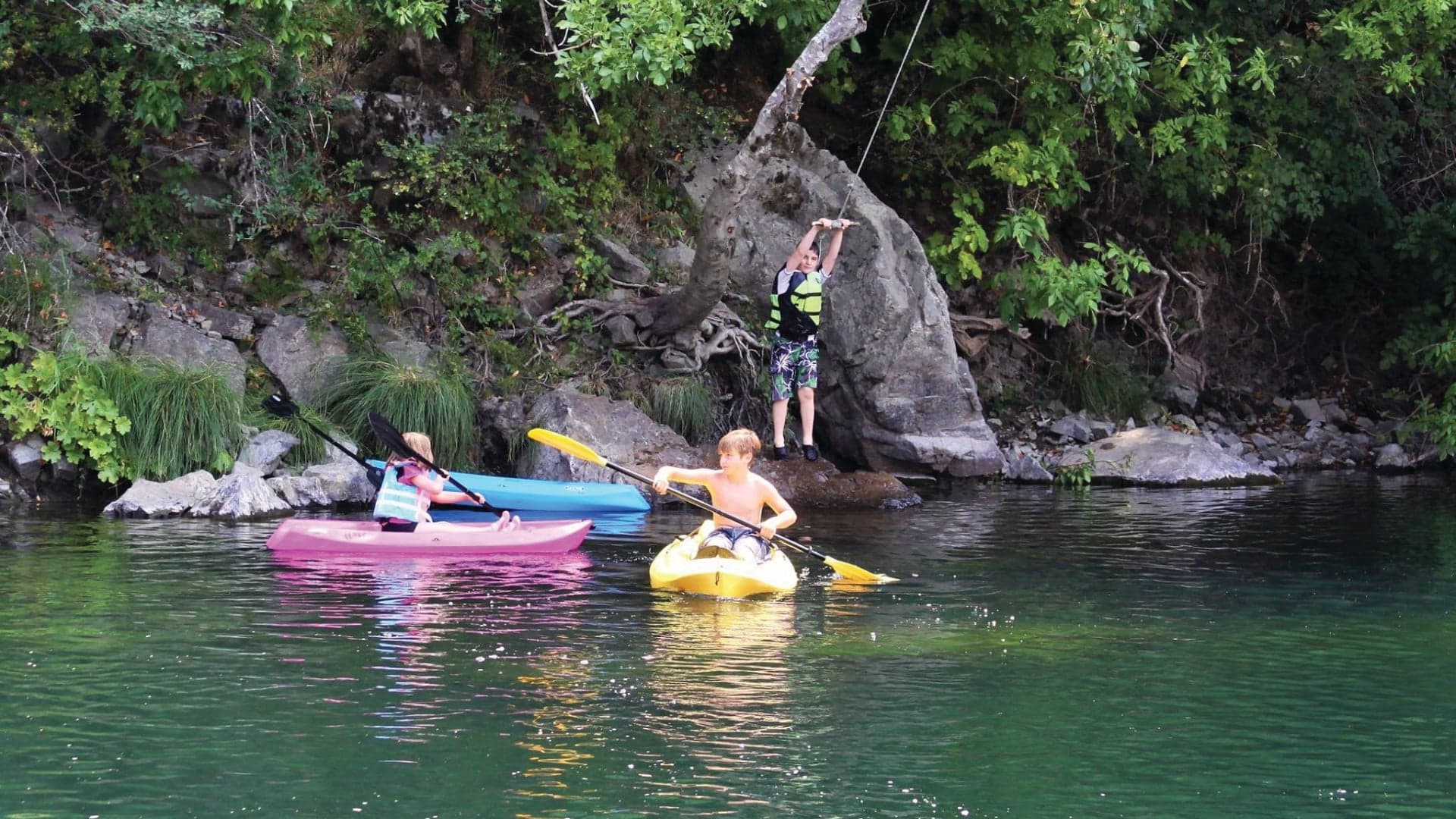 Two children kayaking on a calm water body; one girl in a pink kayak, one boy in a yellow kayak, with a third child on shore.