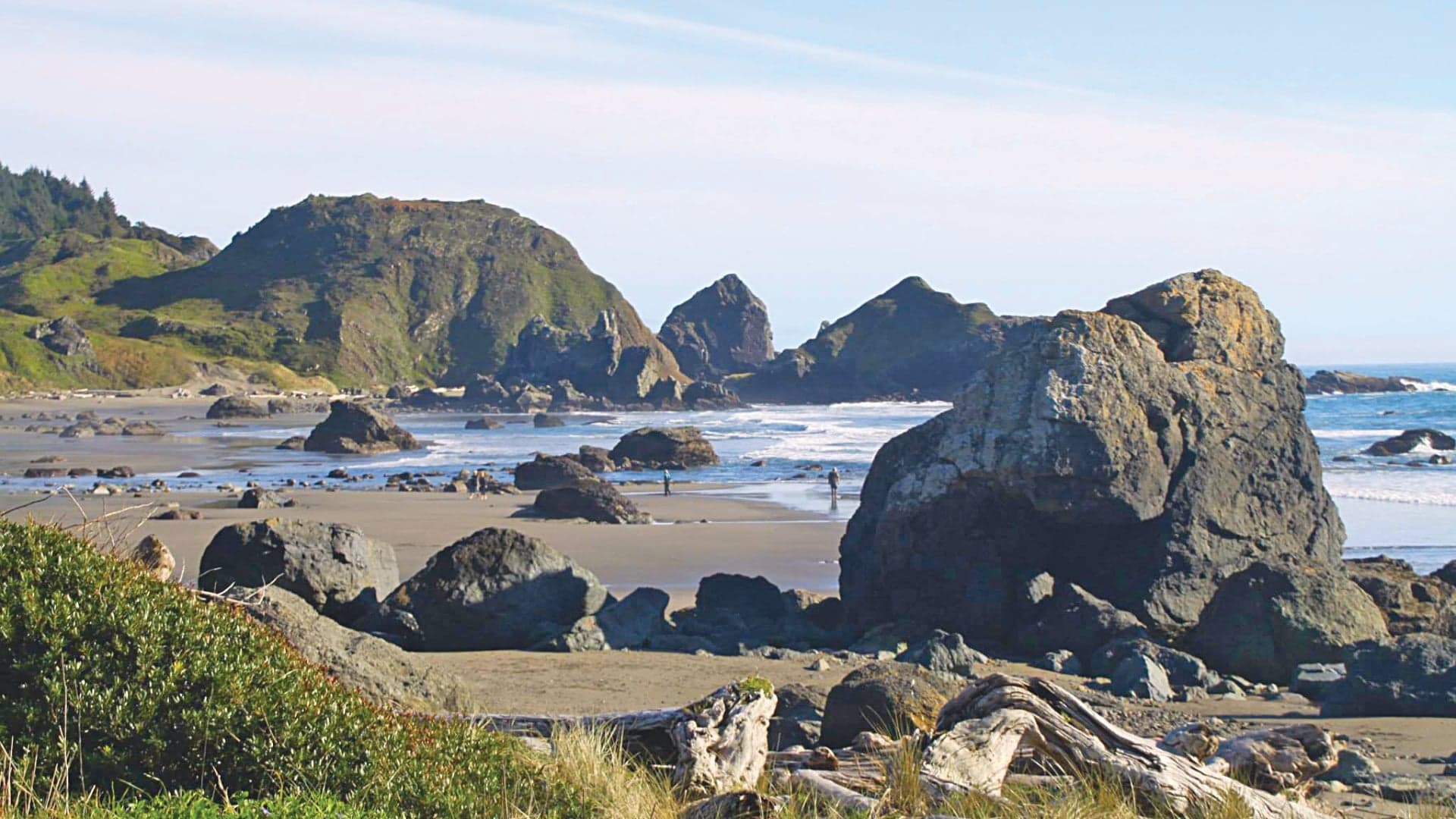 Coastal scene featuring rocky formations, sandy beach, and a calm ocean under a clear blue sky.