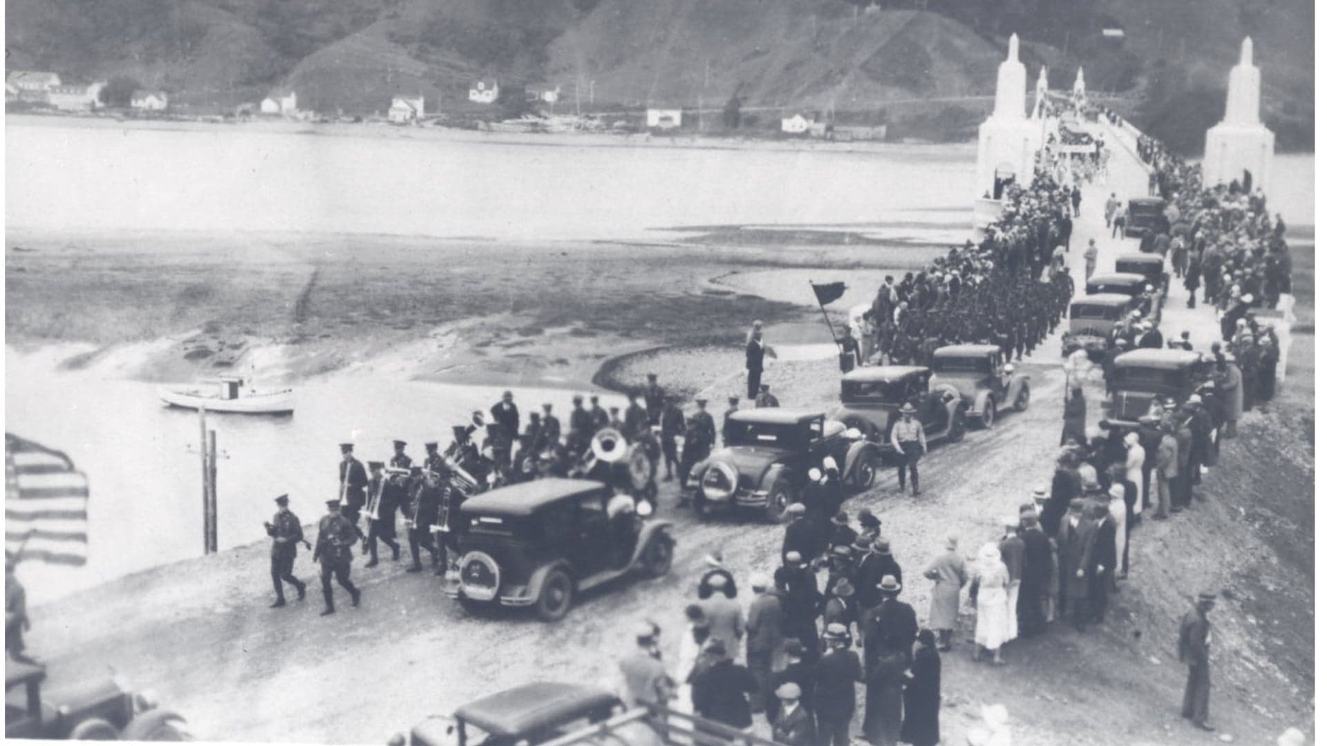 Historic black-and-white photo of a parade with soldiers, cars, and a crowd on a bridge over water.