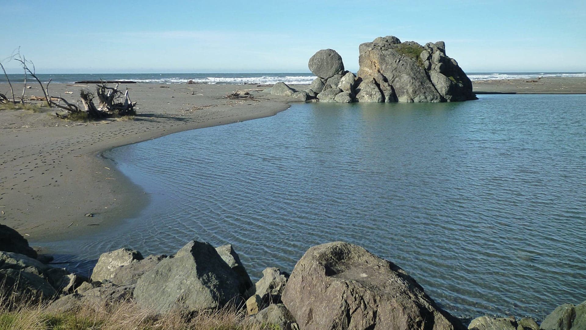 A calm beach scene featuring a rocky outcrop, sandy shoreline, and gentle waves under a clear blue sky.