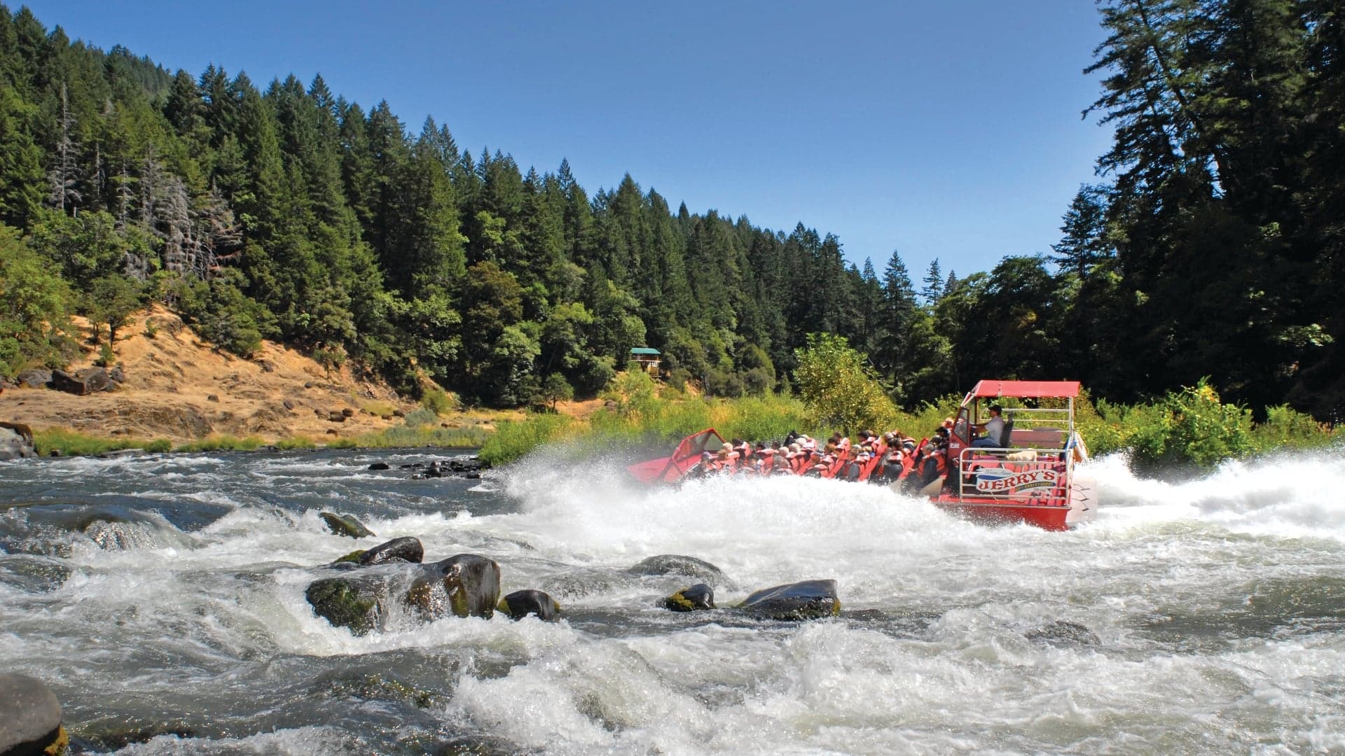A red raft with passengers navigates rapids in a river surrounded by lush green trees and rocky banks.