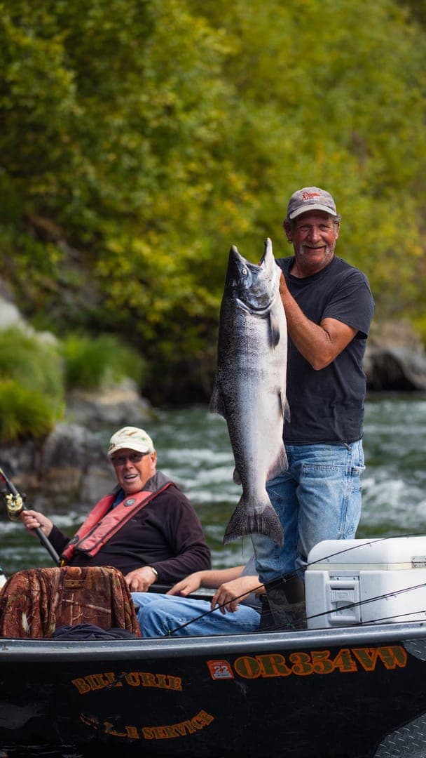 Two men in a boat with a scenic river backdrop; one proudly holds a large fish.