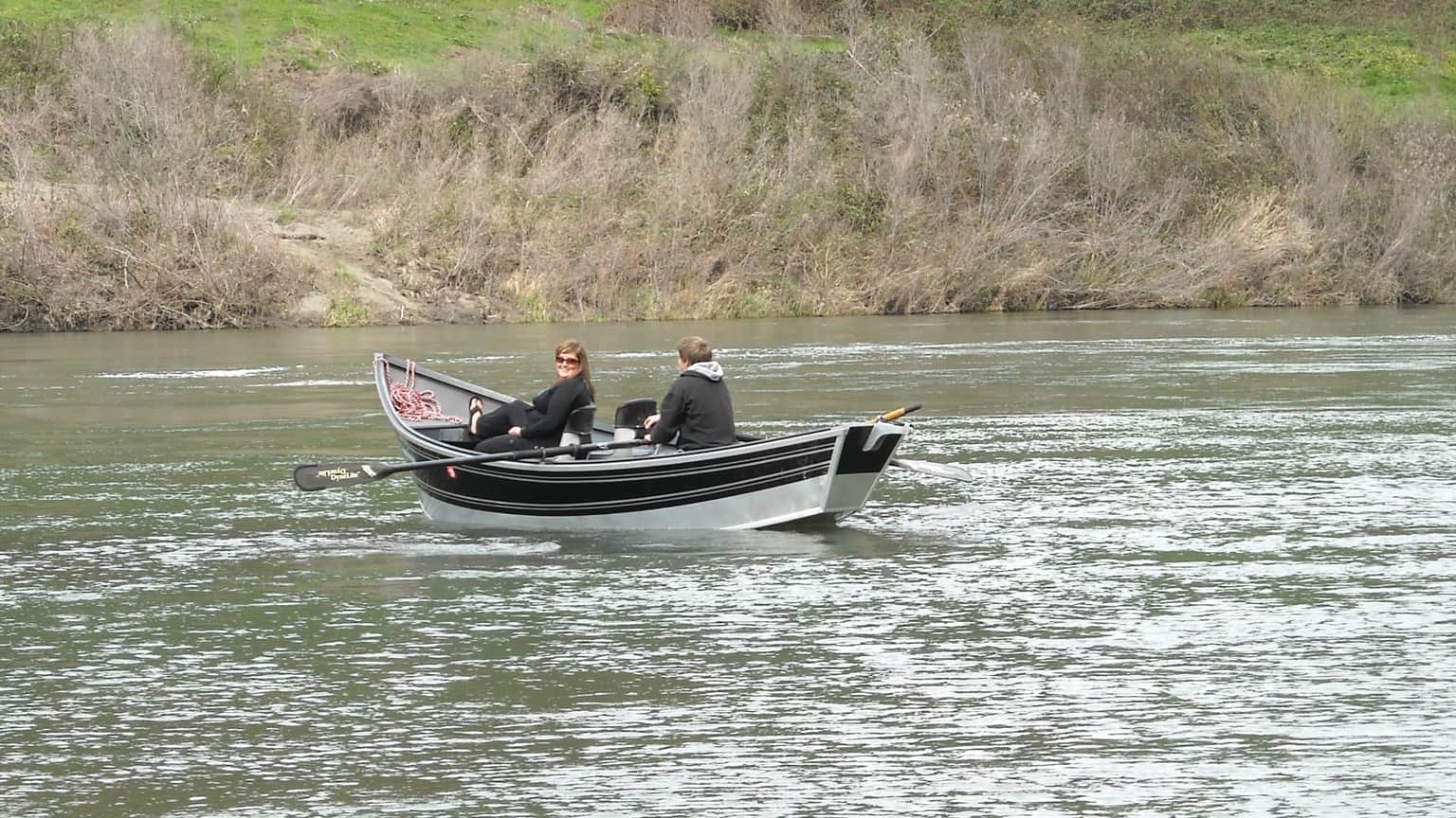 Two people sit in a small boat on a calm river, surrounded by greenery and bare trees.