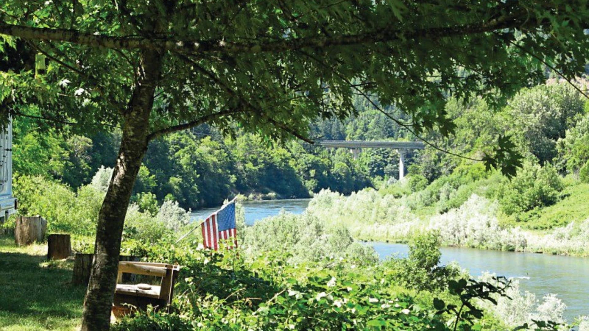 A peaceful riverside view framed by trees, featuring a visible American flag and a bridge in the background.