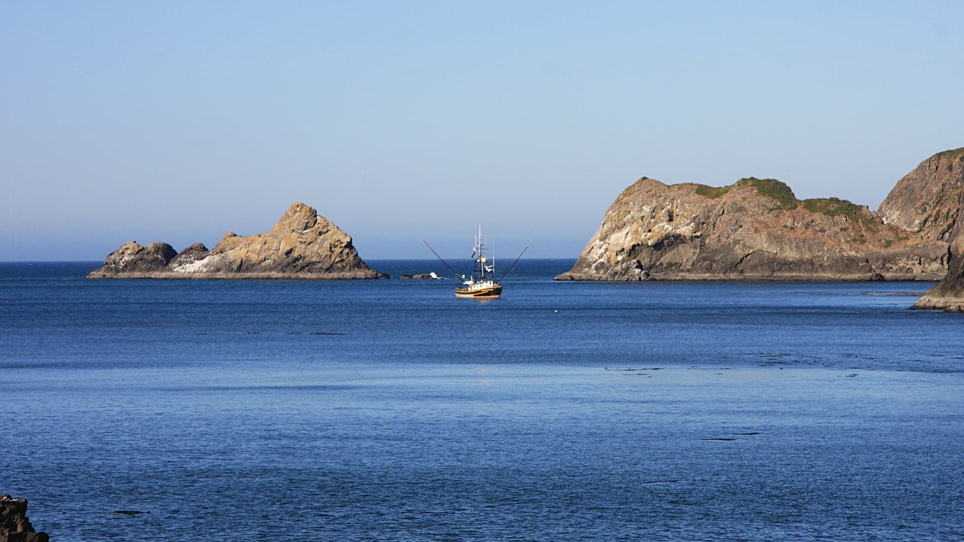 A fishing boat floats in calm blue water between rocky outcrops under a clear sky.