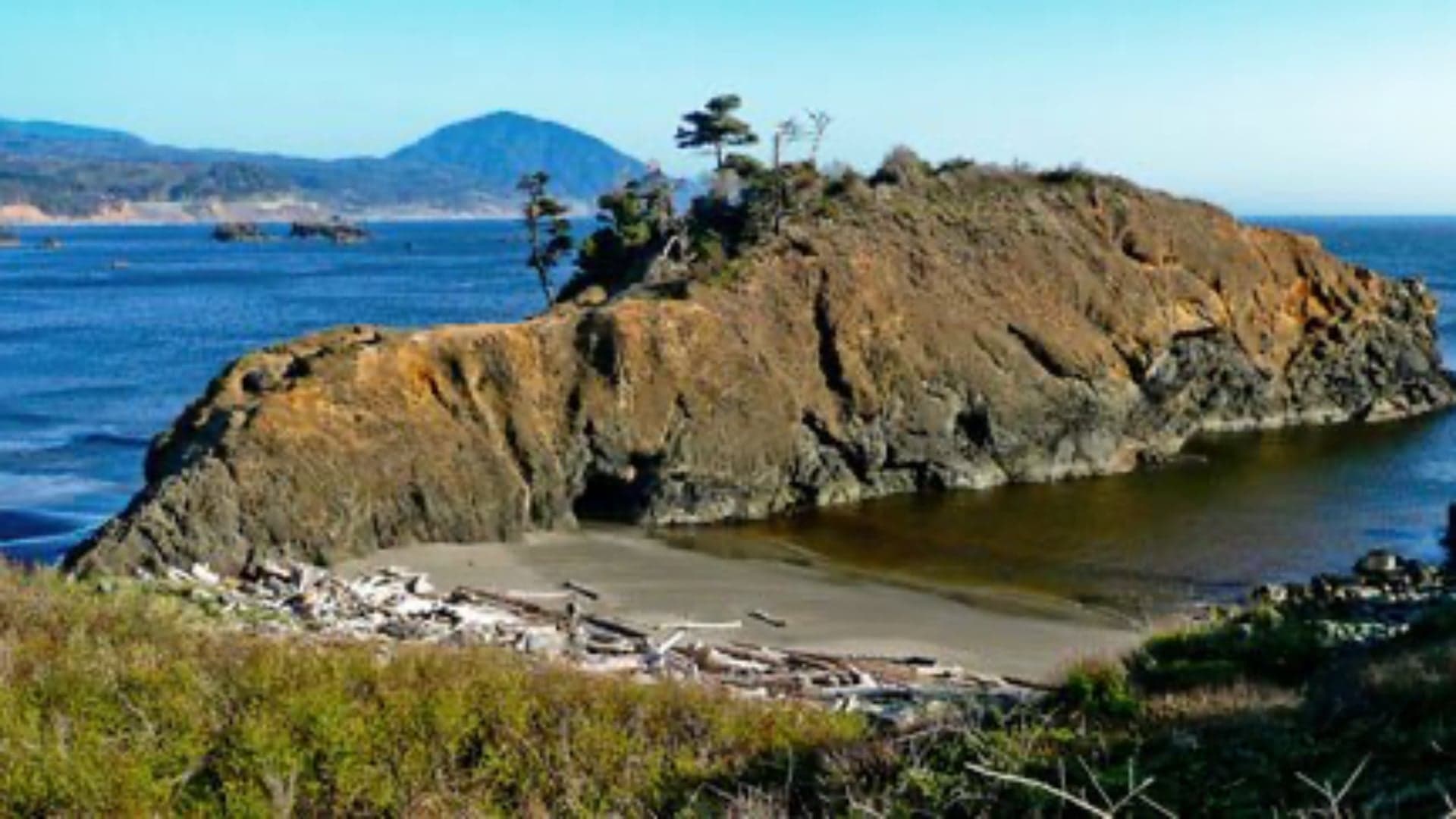 Rocky coastal landscape with vegetation, a sandy beach, and calm water under a clear blue sky.