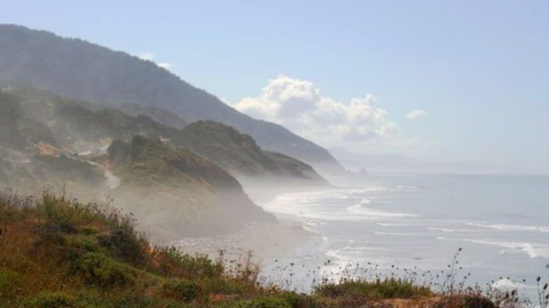 Coastal view with mountains and mist, waves gently hitting the shore under a blue sky.