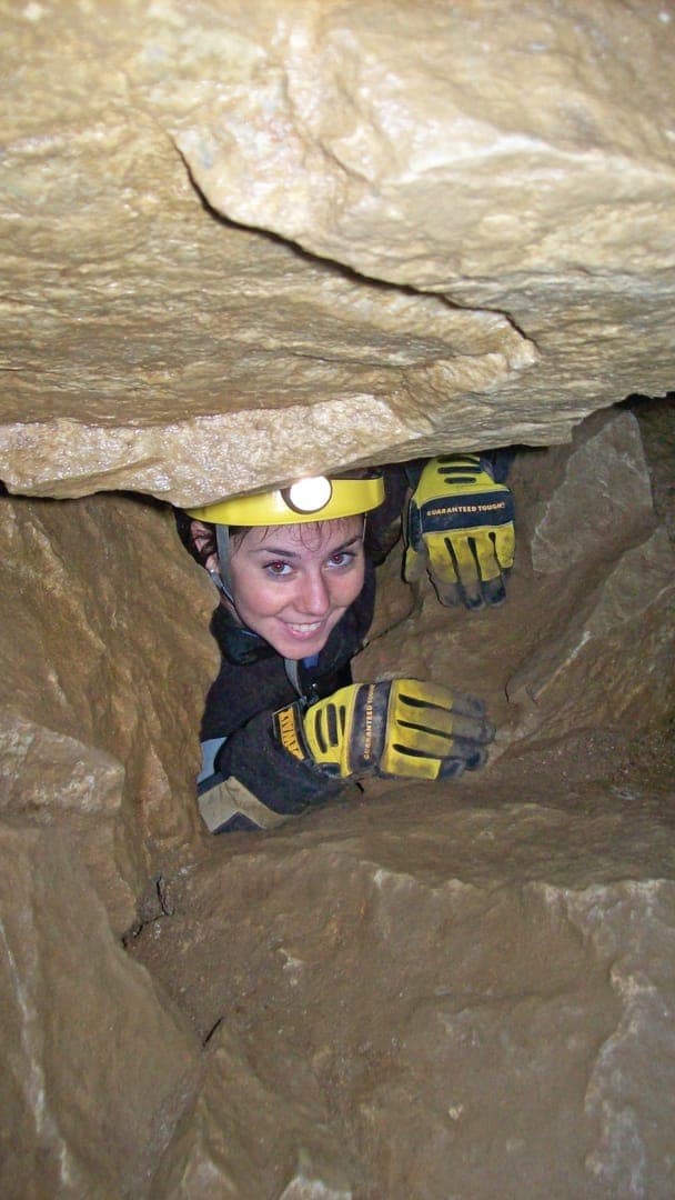 A person in a helmet smiles while crawling through a narrow cave passage, wearing gloves and a dark outfit.