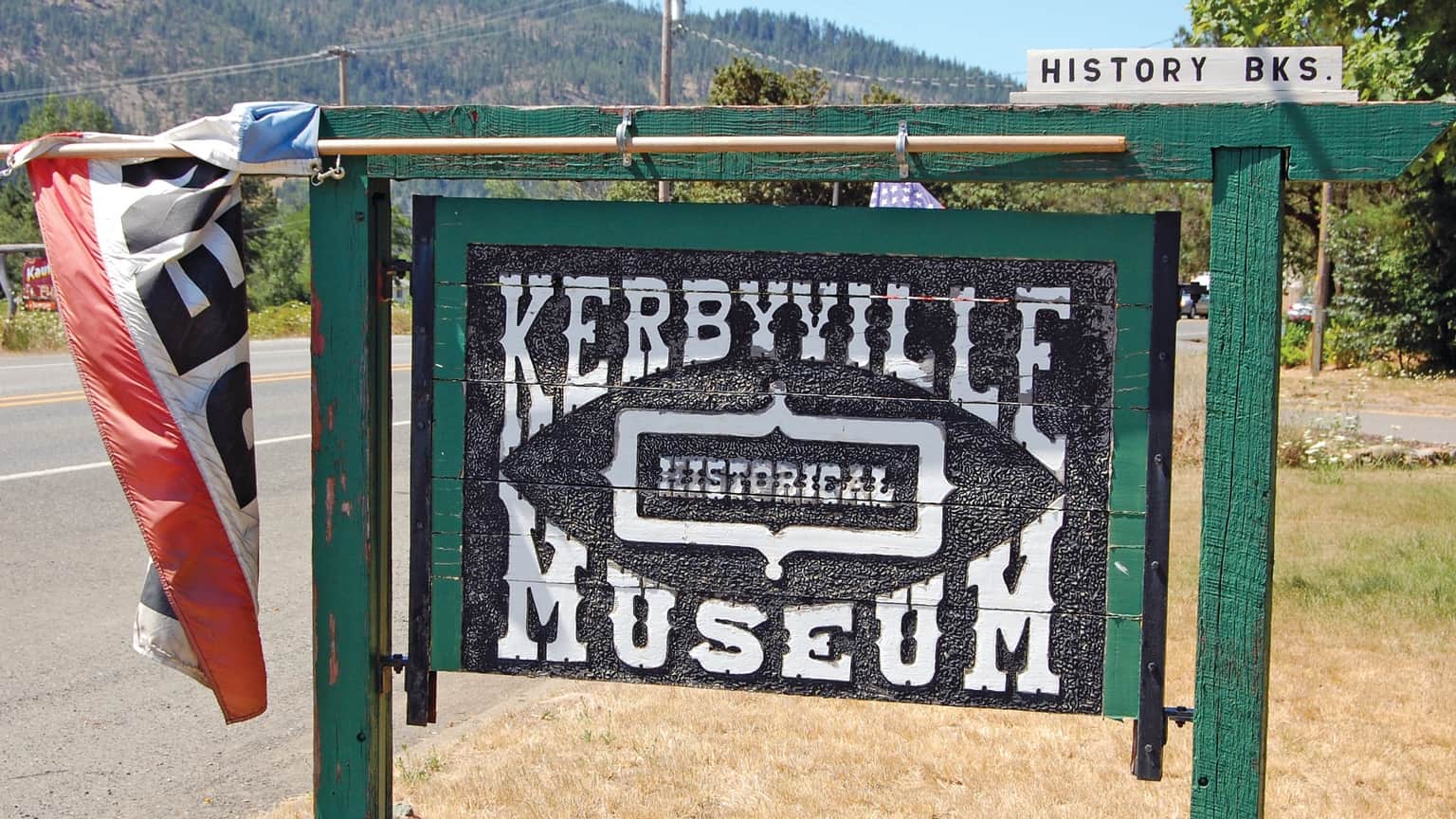 Sign for Kerbyville Historical Museum, with red and black banner and green frame, by a road.