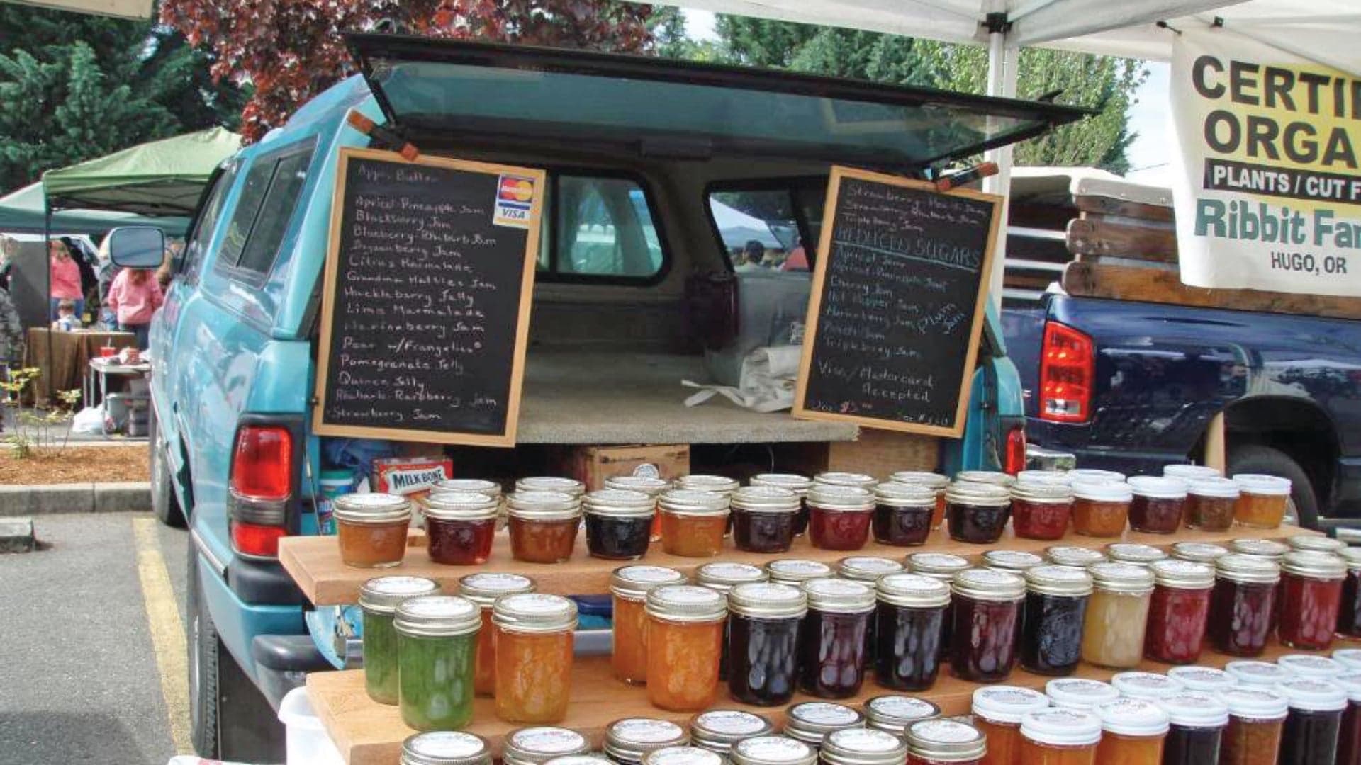 A blue truck offers jars of colorful jams on a wooden stand under a market tent, with chalkboard menus displayed.