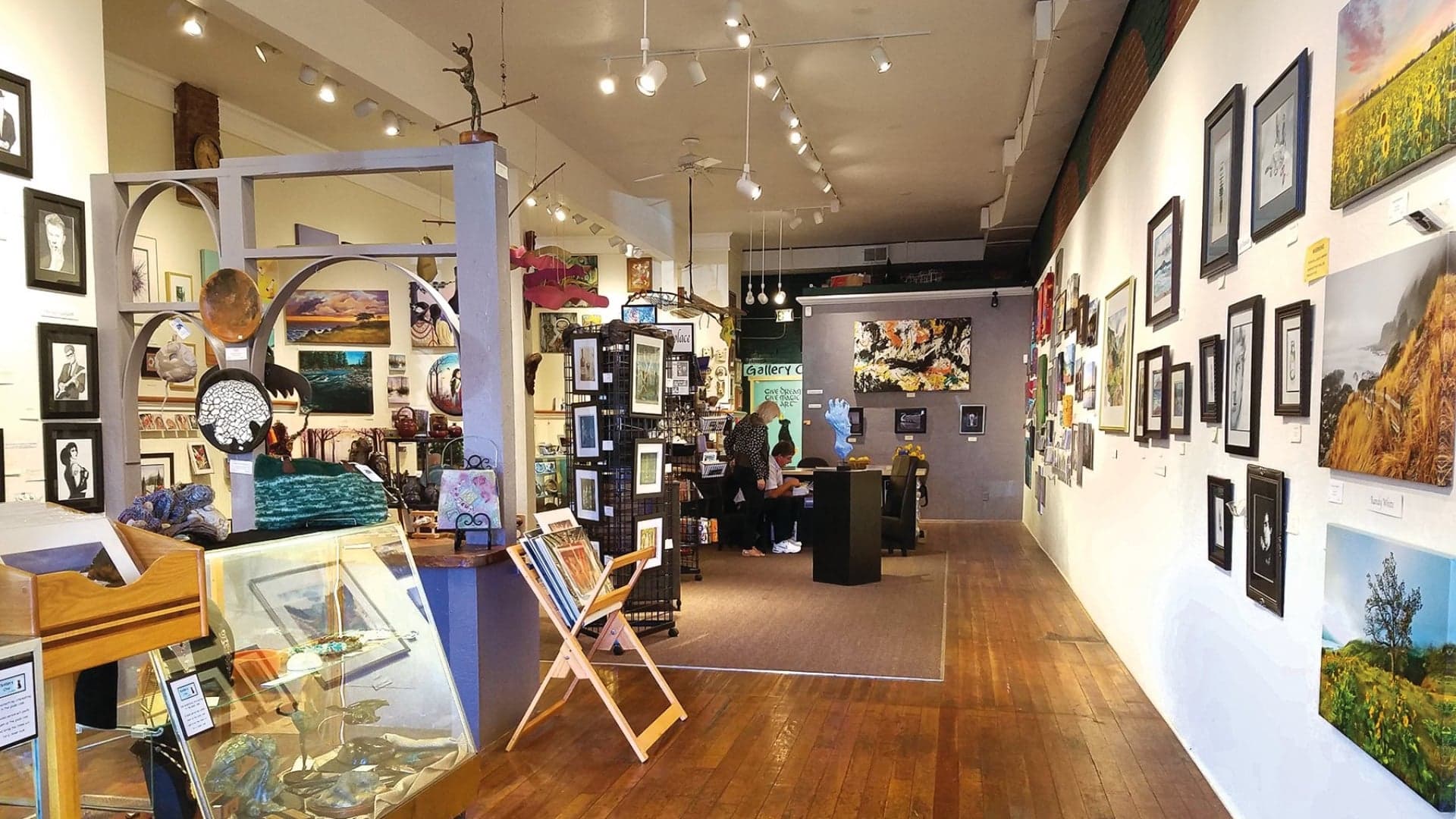 Interior of an art gallery featuring various artworks, sculptures, and display stands with a visitor at the counter.