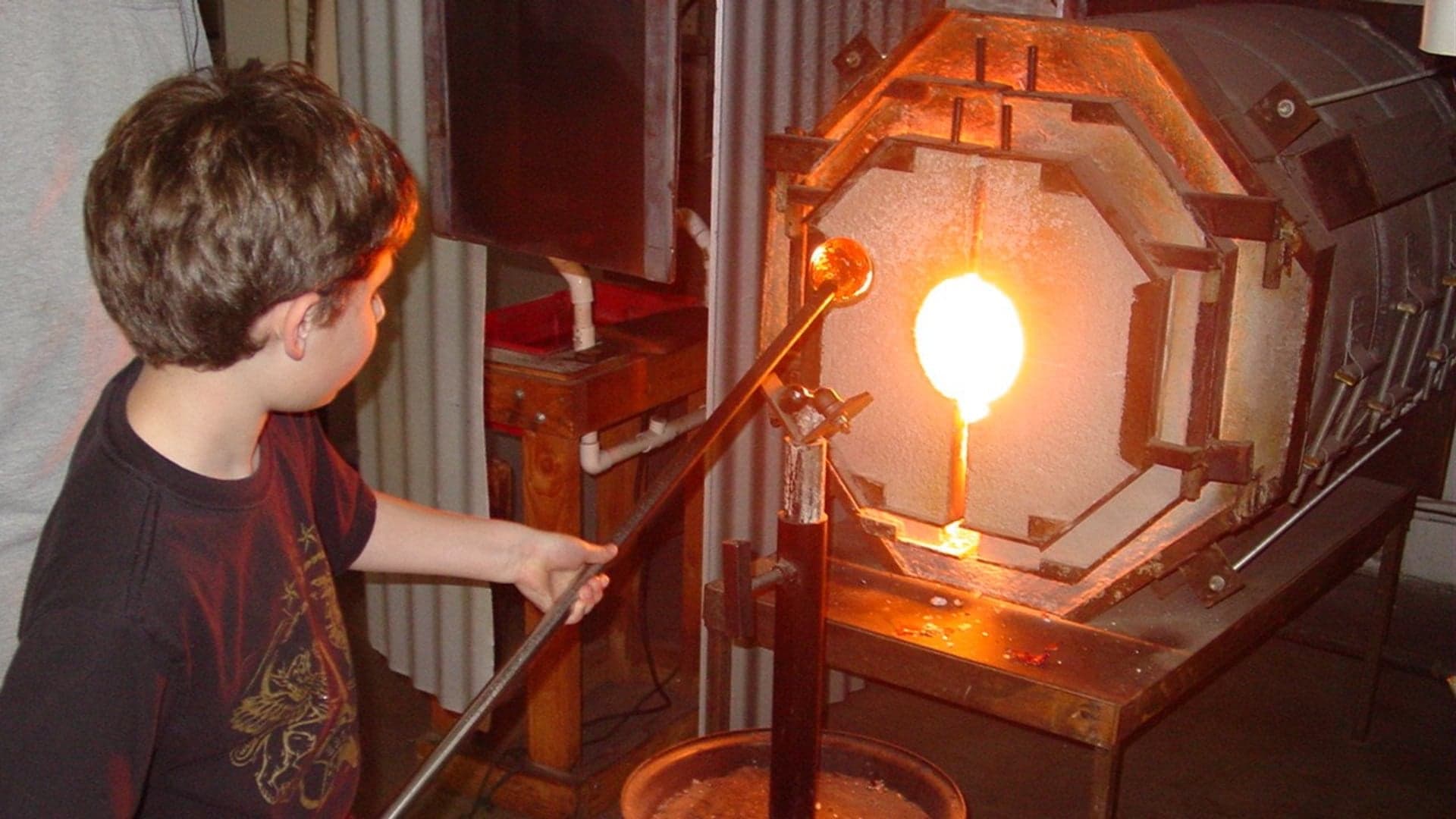 A child observes and uses a blowpipe to shape molten glass in a glassblowing studio.