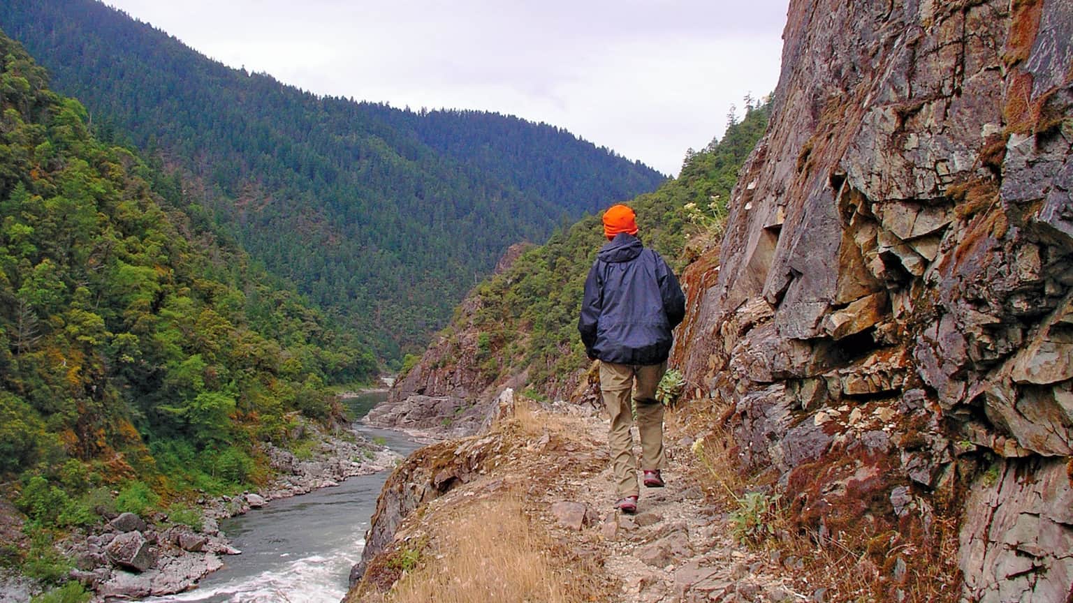 A hiker wearing an orange hat walks along a rocky path beside a river, surrounded by lush green mountains.