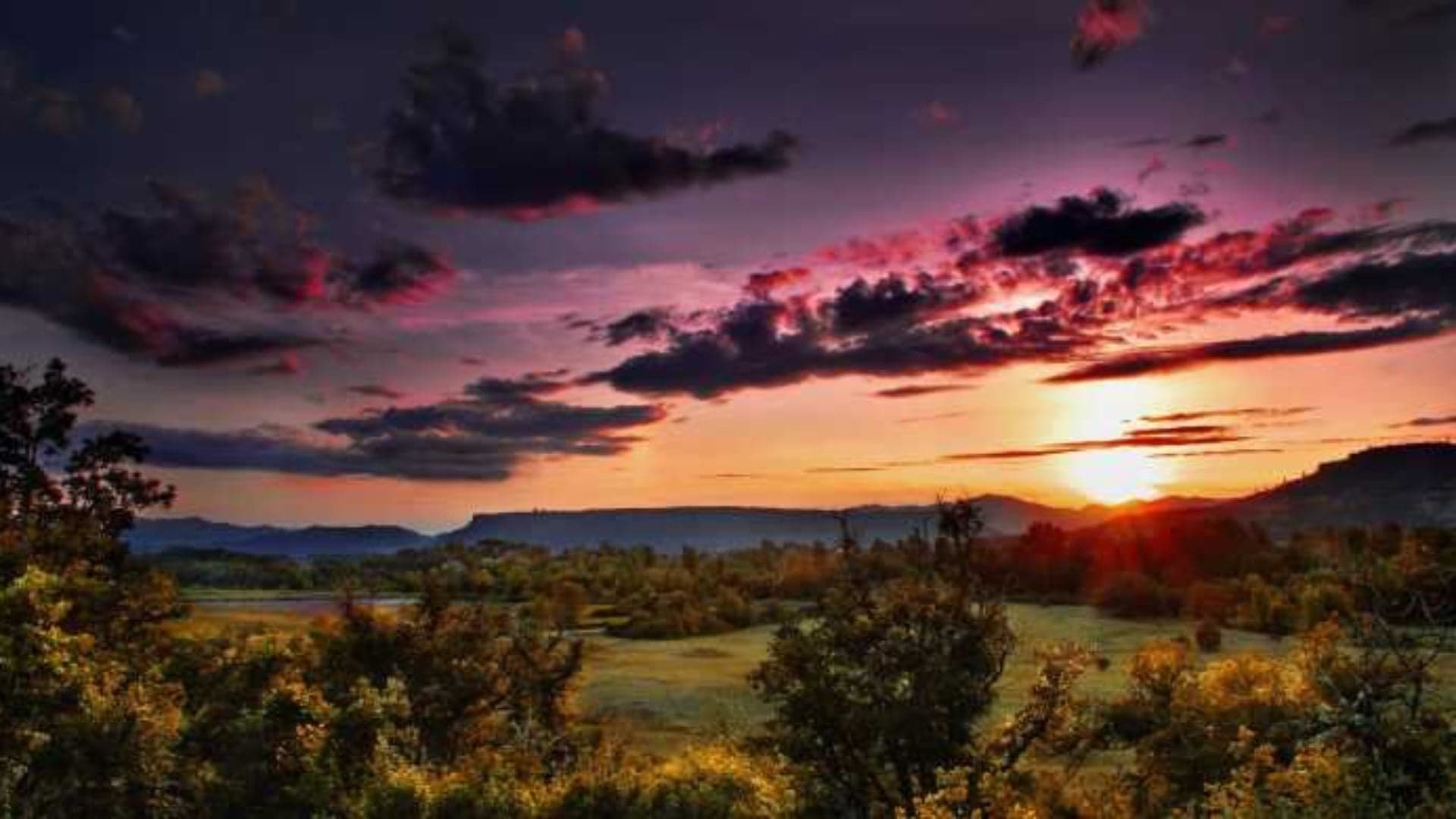 Vibrant sunset over rolling hills, with colorful clouds and foreground trees.