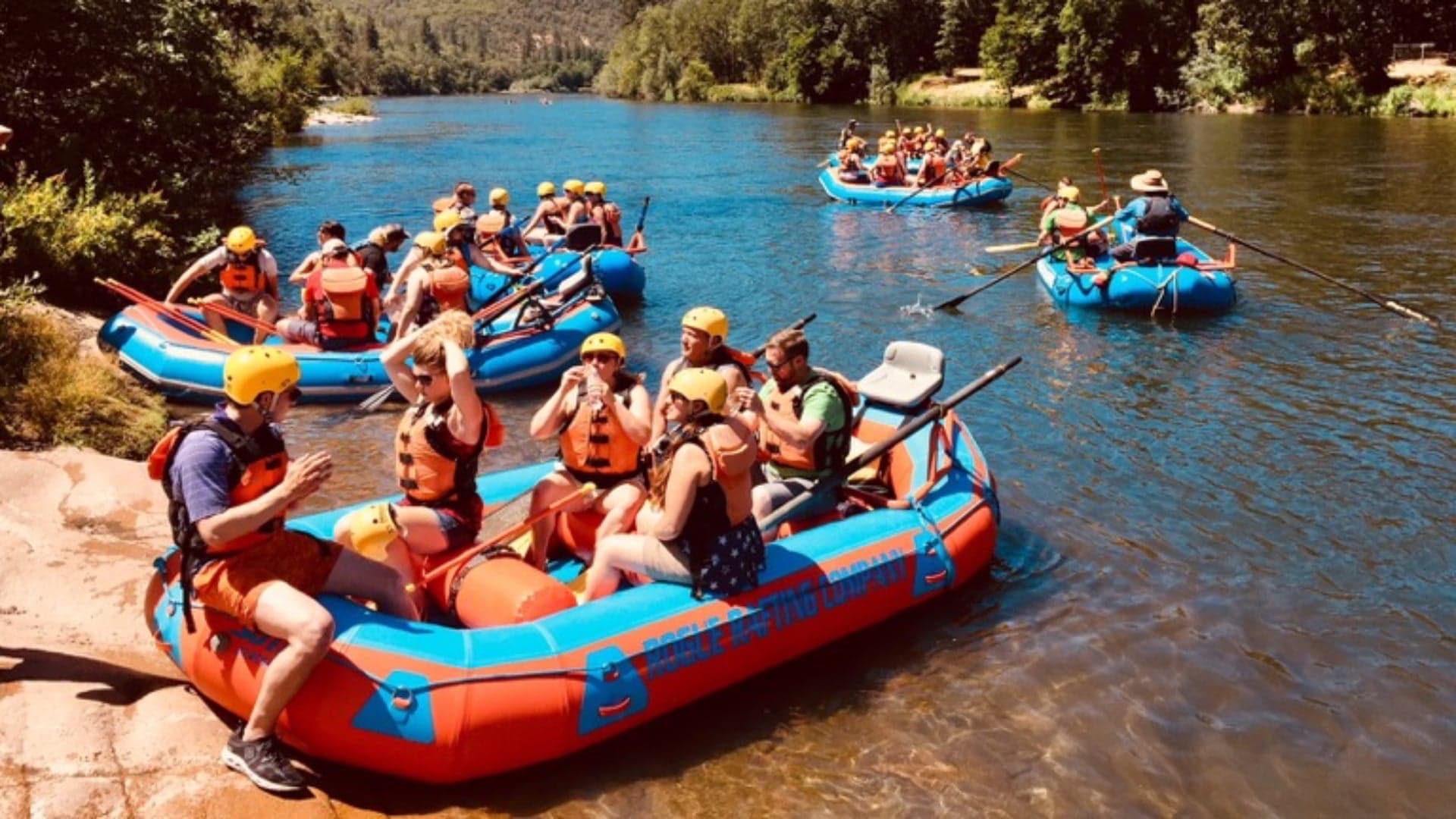 A group of people in raft boats on a river, preparing for whitewater rafting with life jackets and helmets.