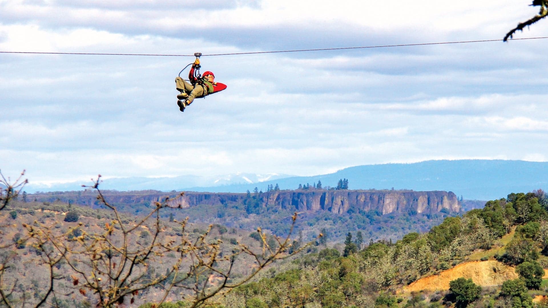 A person zip-lining across a scenic landscape with mountains and trees in the background.
