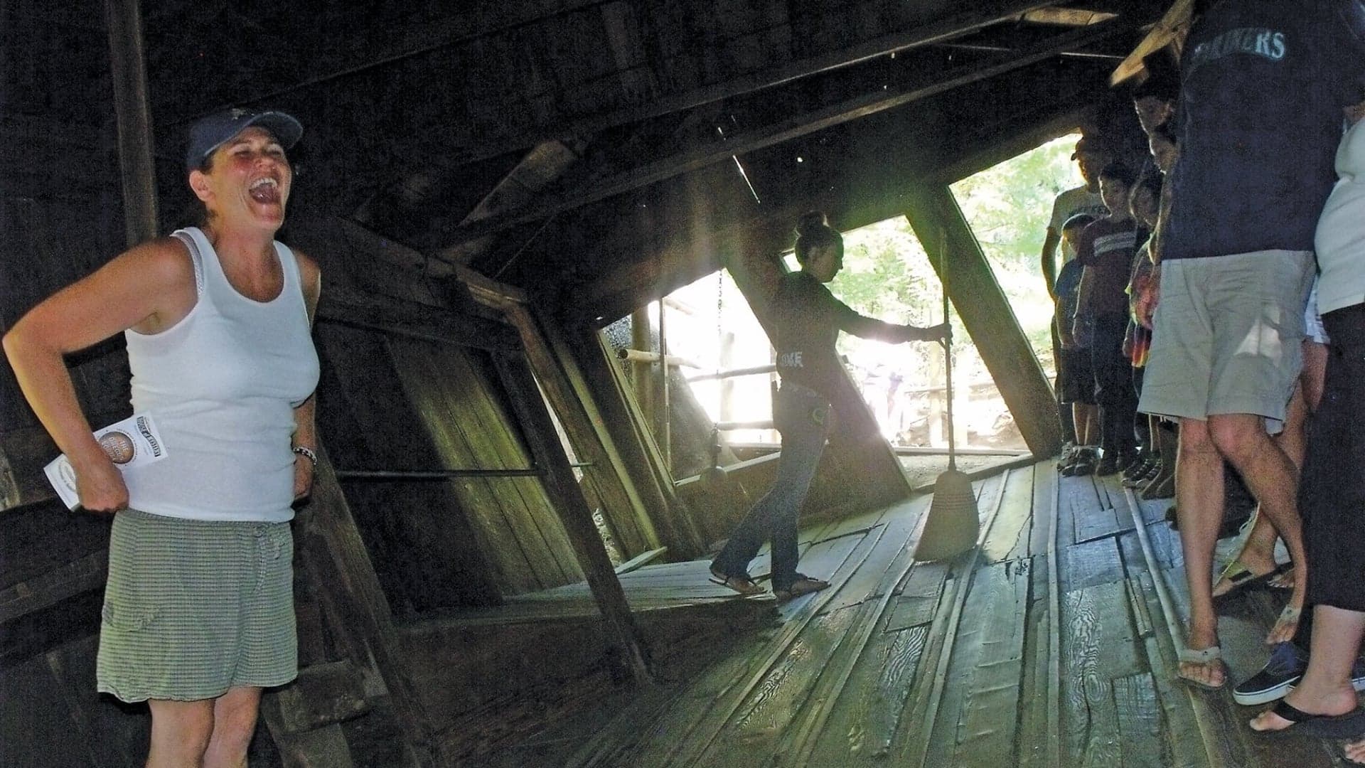 A woman laughs while standing inside a slanted wooden structure, with a small group watching nearby.