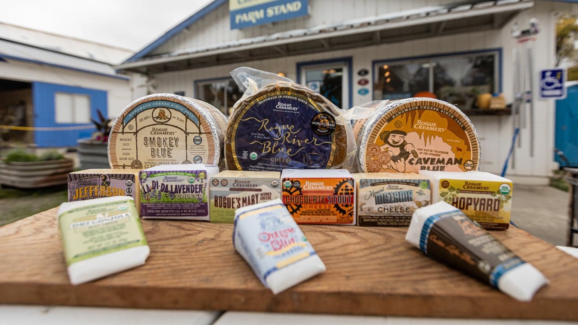 Various cheeses from Rogue Creamery displayed on a wooden board outside a farm stand.