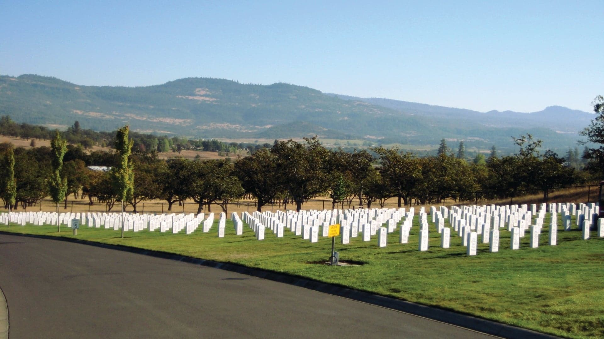 A serene cemetery with rows of white gravestones, surrounded by trees and mountains in the background.