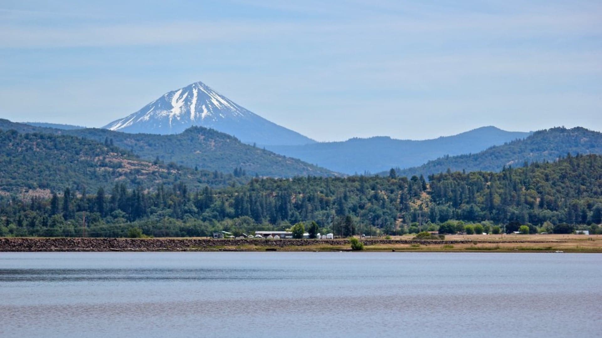 Snow-capped mountain rises behind green hills and a calm lake under a clear sky.