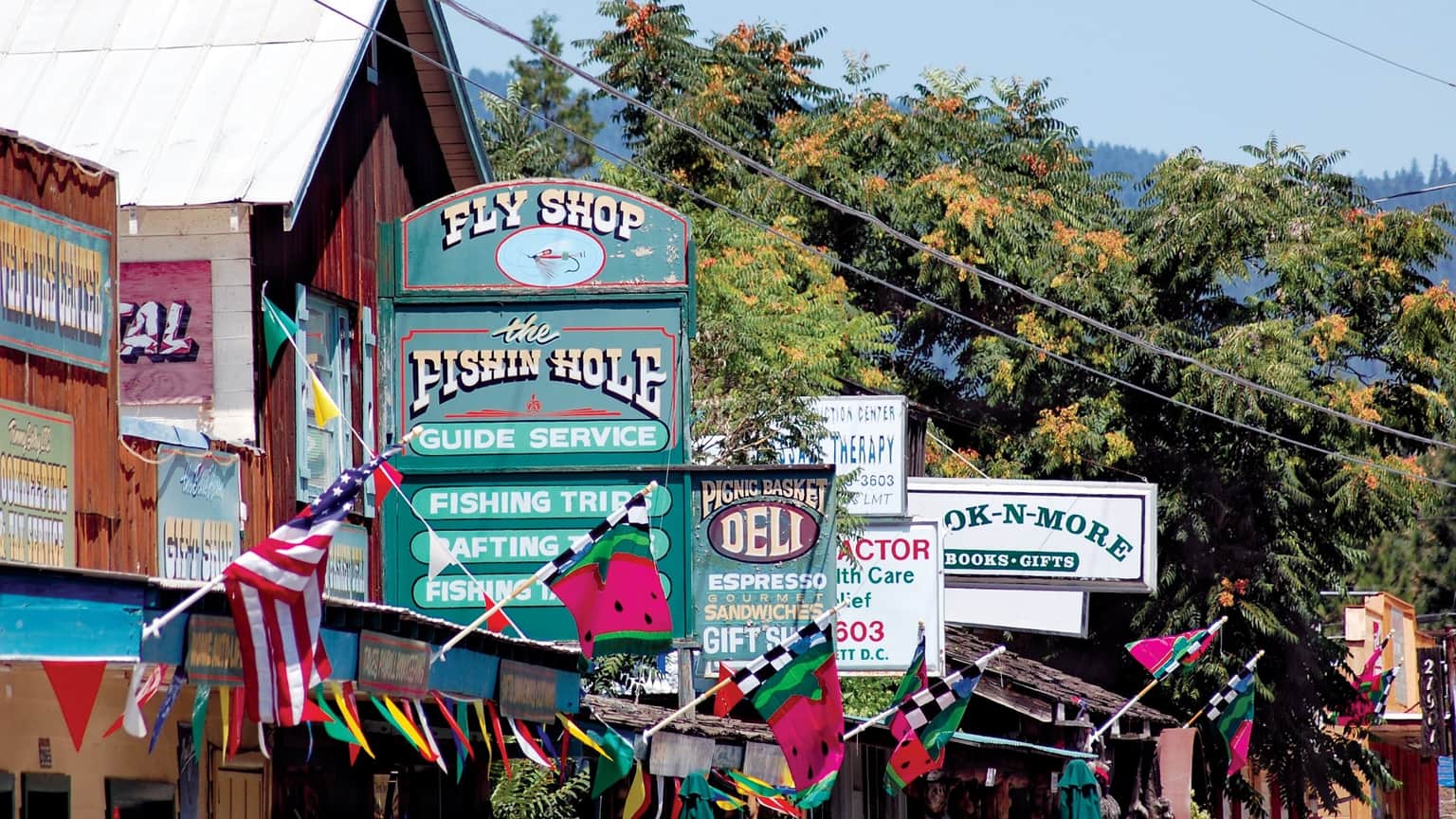 Colorful storefronts and signs lining a street, featuring a fly shop, deli, and various shops with flags.