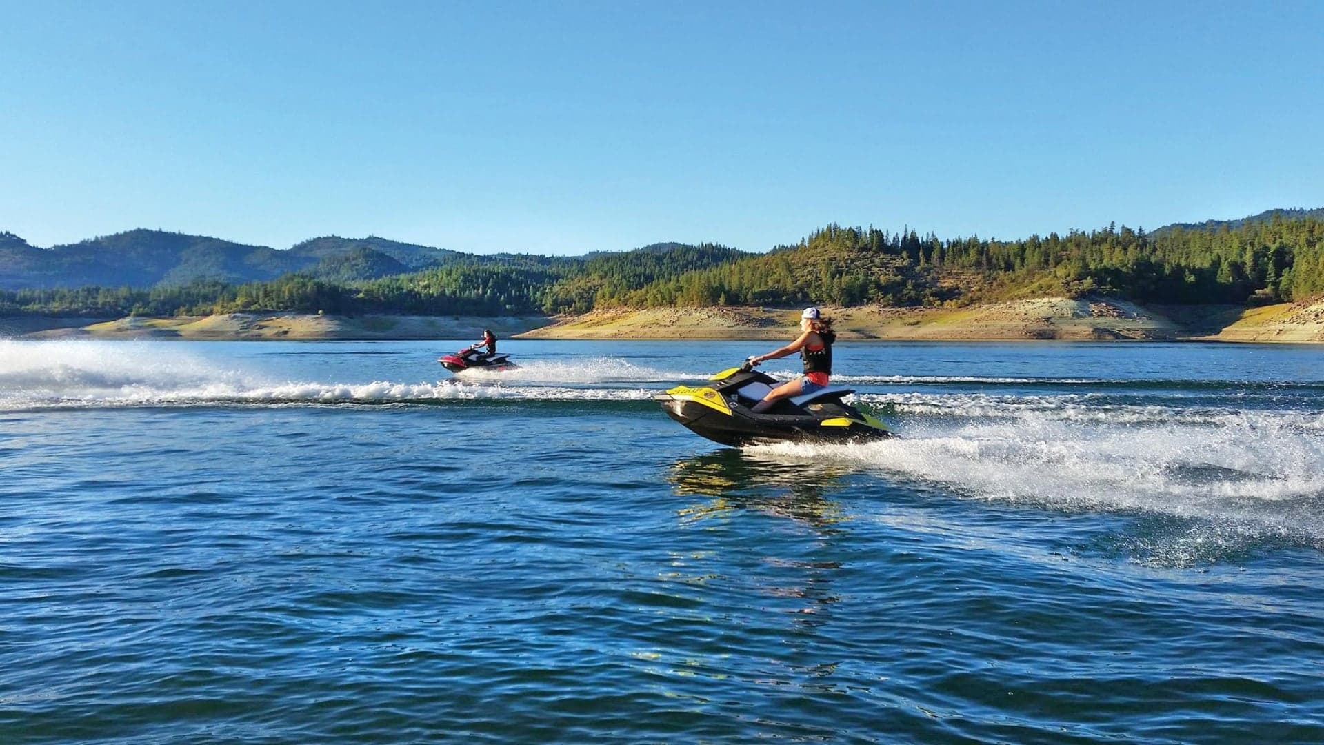 Two people riding jet skis on a lake, surrounded by hills and trees under a clear blue sky.