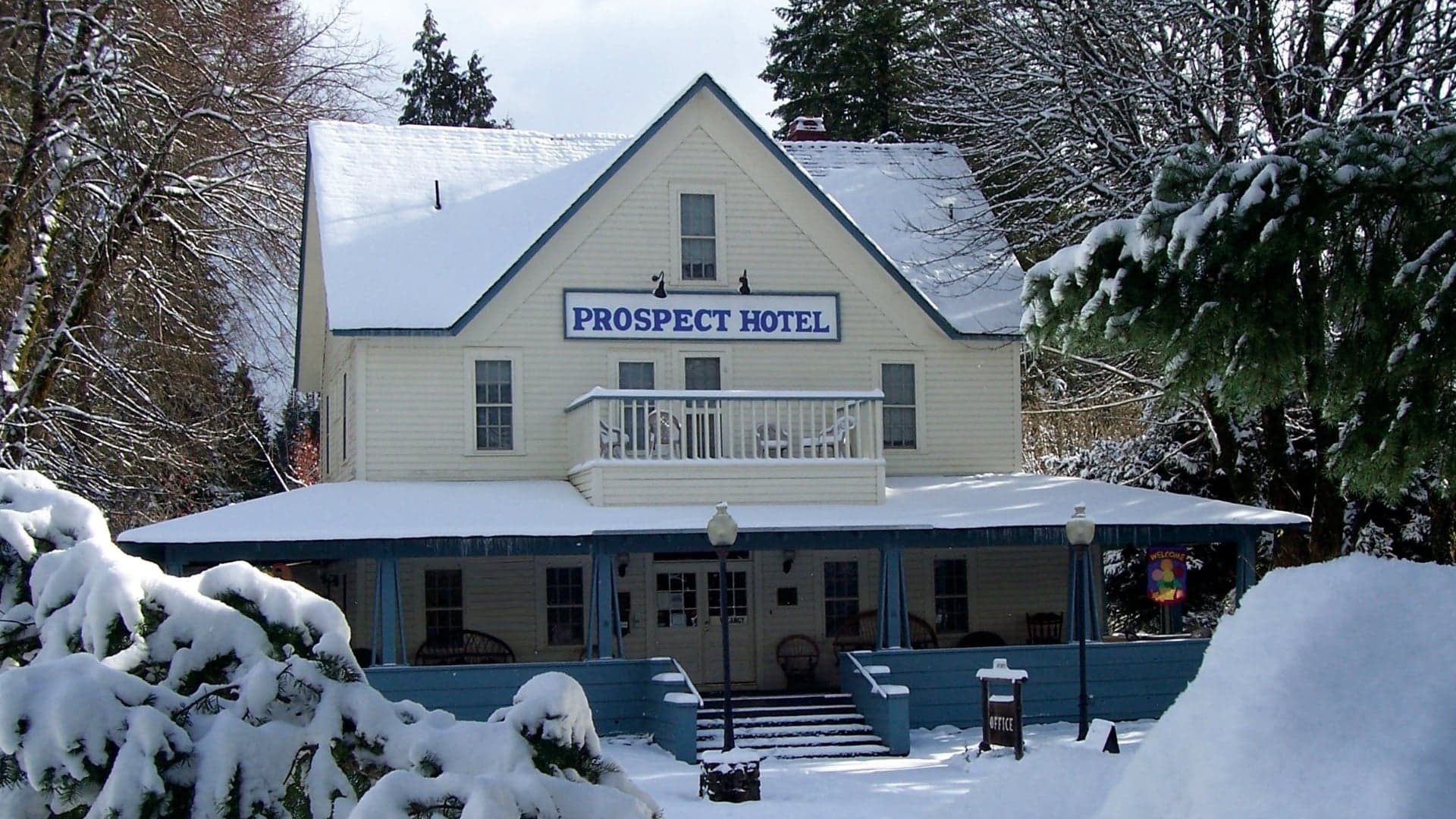 The Prospect Hotel is covered in snow, featuring a blue porch and large sign. Surrounding trees are snow-laden.