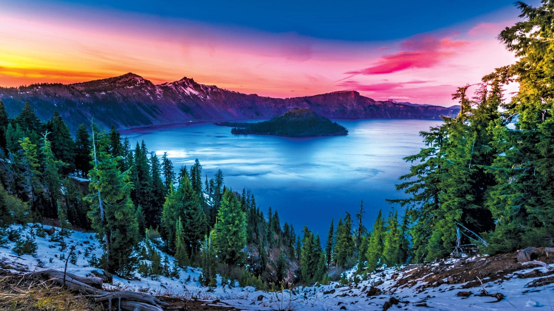 Sunset over Crater Lake, with an island in the center and lush green trees in the foreground.