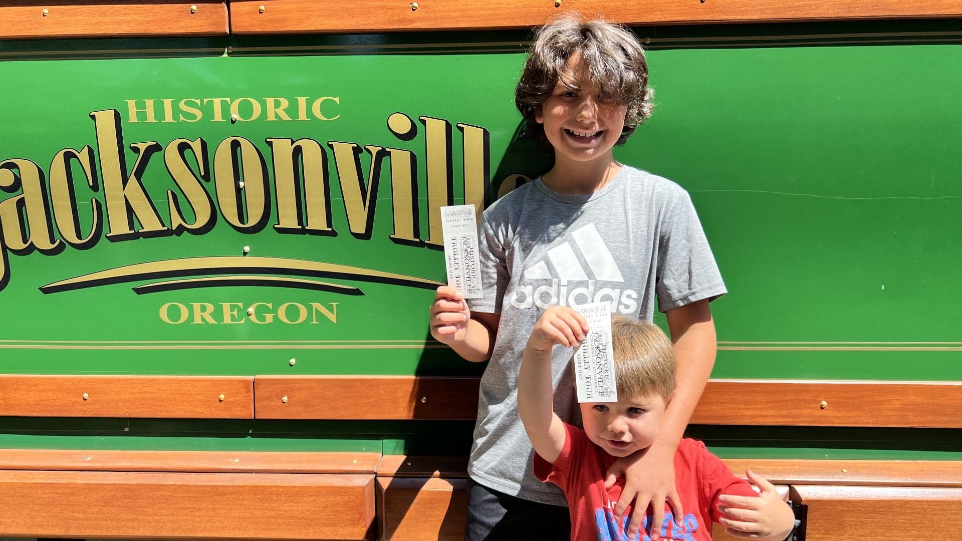 Two children stand in front of a green sign that reads "Historic Jacksonville, Oregon," each holding tickets.
