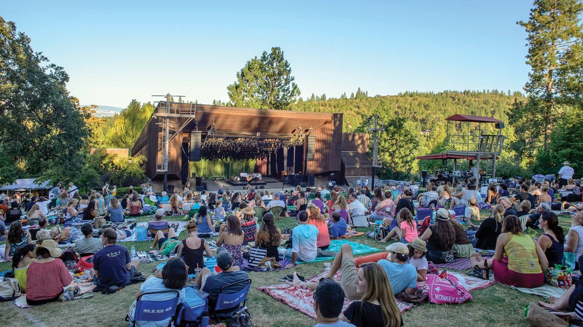 A large audience sits on grassy hillsides at an outdoor concert venue surrounded by trees.
