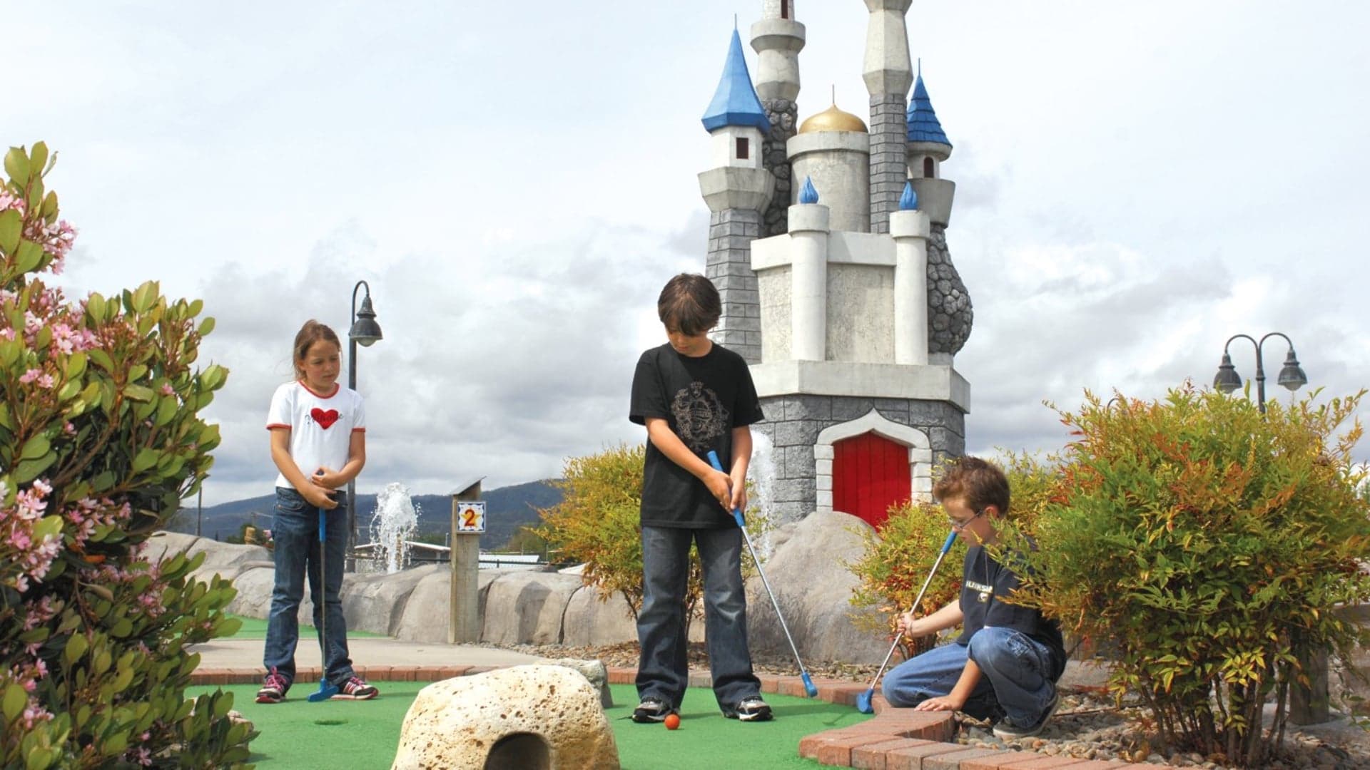 Three children play miniature golf in front of a castle-themed structure on a cloudy day.