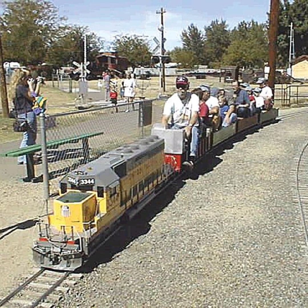 A miniature train with passengers curves along a track in a park, surrounded by trees and spectators.
