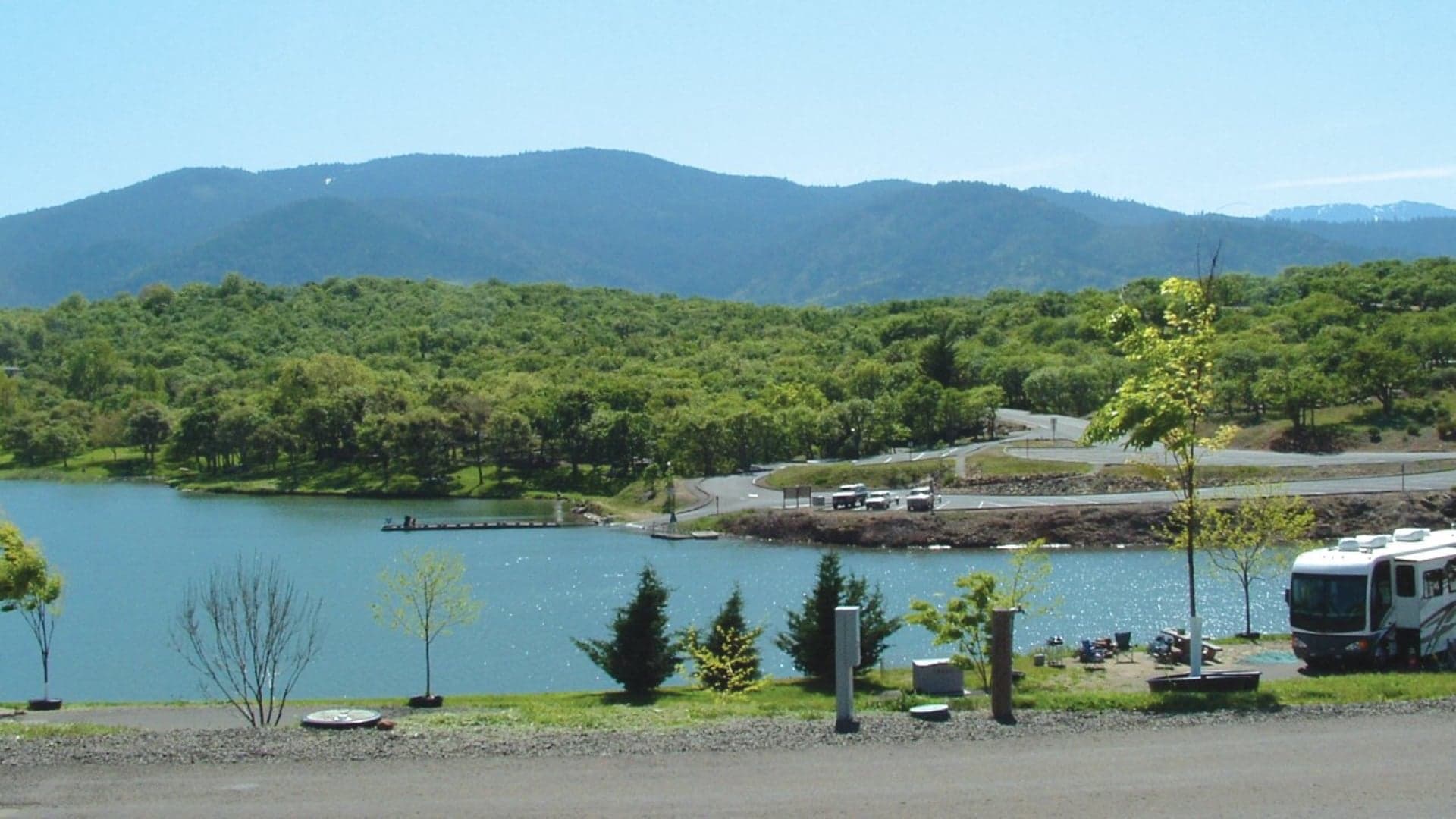 Scenic view of a calm lake surrounded by green hills, with a roadway and parked vehicles visible nearby.