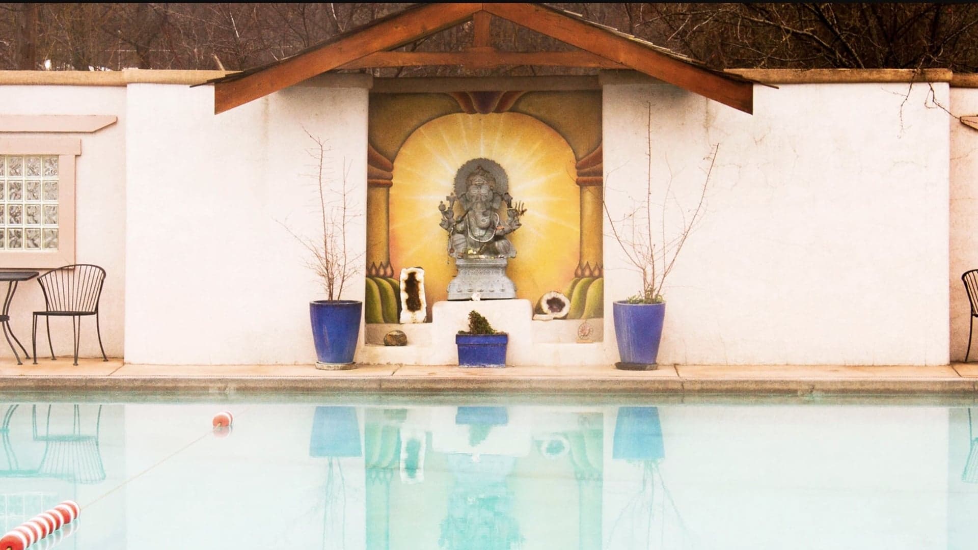 A tranquil pool scene featuring a statue of a deity against a wall, surrounded by blue planters and a chair.