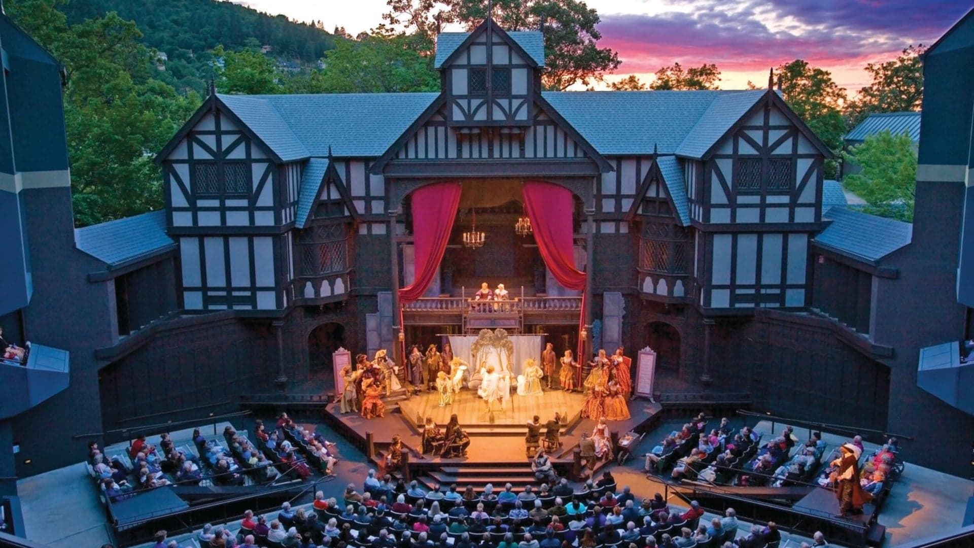 A theater performance on a stage with an ornate backdrop, surrounded by an audience in an outdoor setting at sunset.