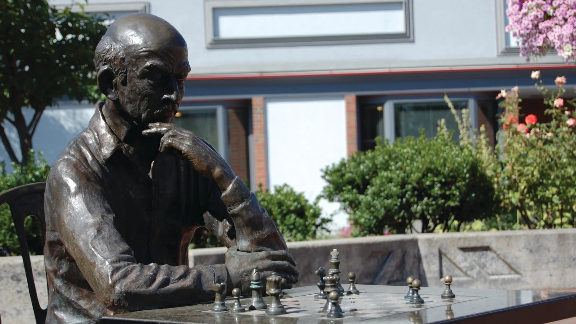 Bronze statue of a man deep in thought while playing chess outdoors, surrounded by greenery and flowers.