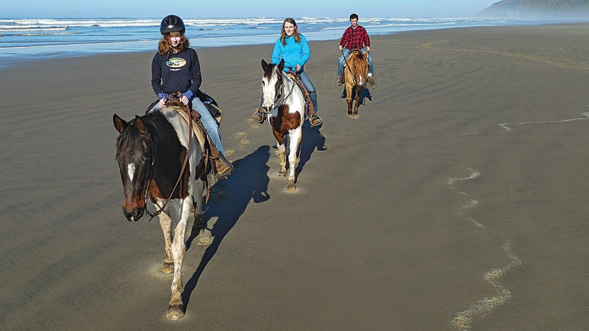 Three riders on horseback trotting along a sandy beach with ocean waves in the background.